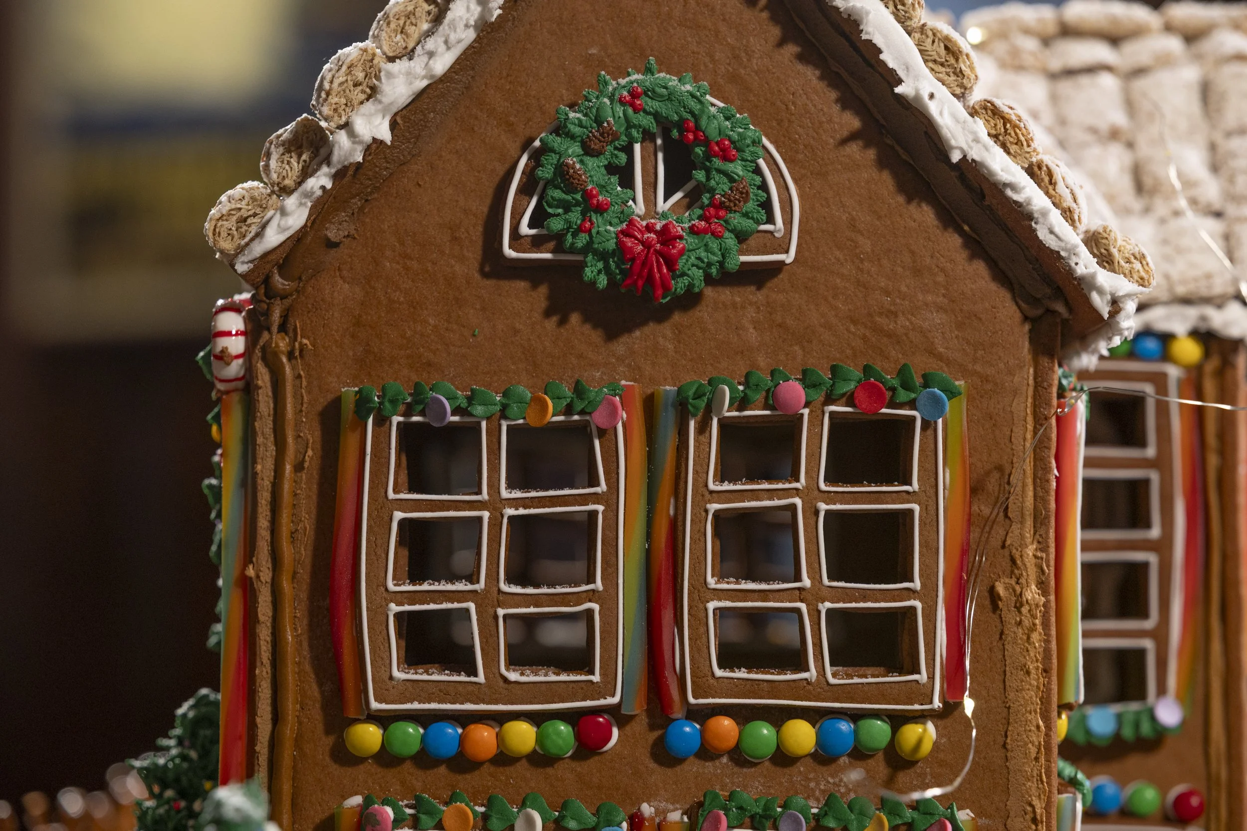Decorated gingerbread house with windows outlined in white icing, green piped icing trim, and colorful candy accents, topped with a Christmas wreath, with another gingerbread house partially visible in the background.