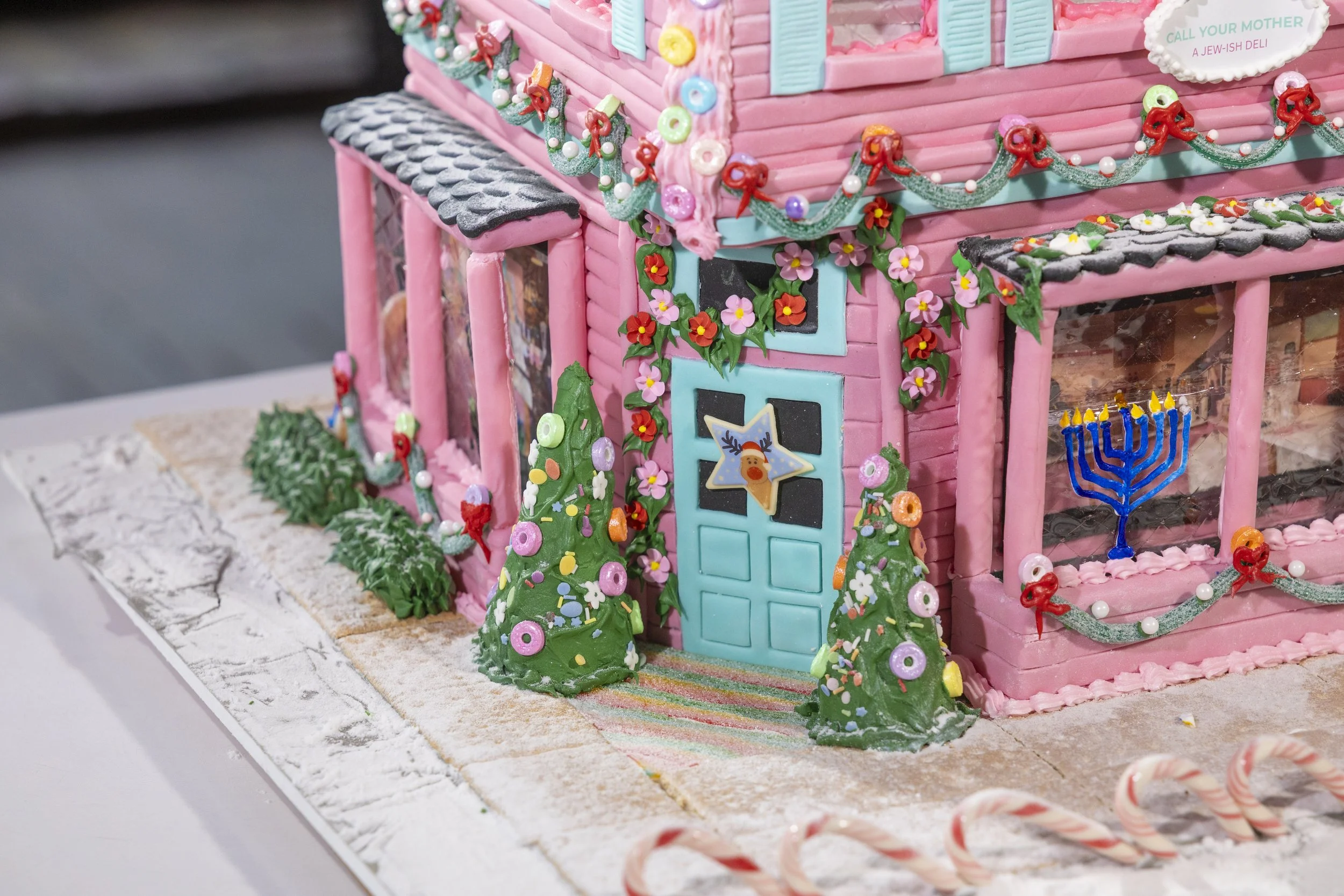 Decorated gingerbread house with pink walls, teal door, Christmas trees, candy canes strung with red bows, and festive ornaments.