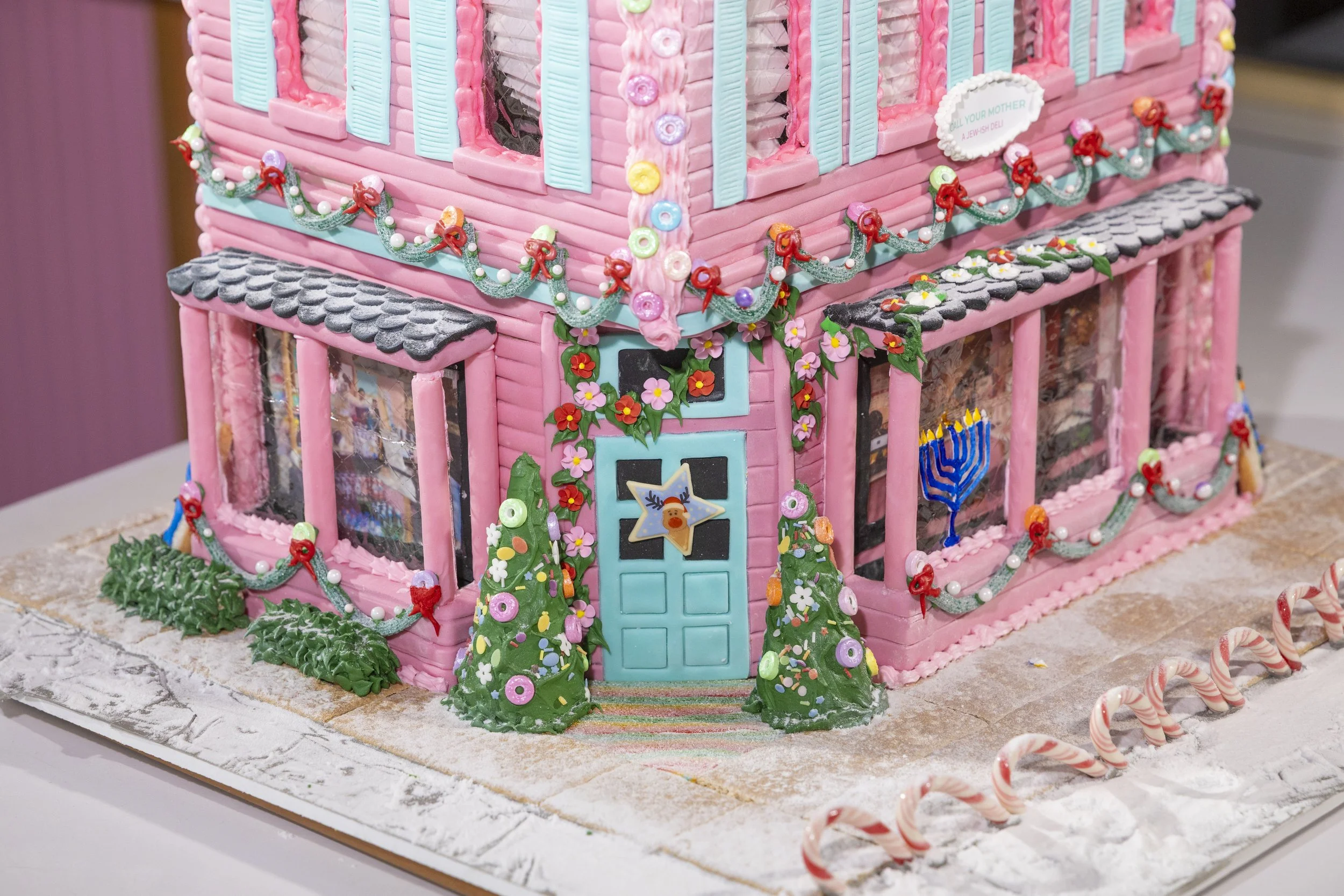 Decorated gingerbread house with pink walls, blue shutters, and Christmas decorations, including garlands, Christmas trees, and holiday-themed figurines.