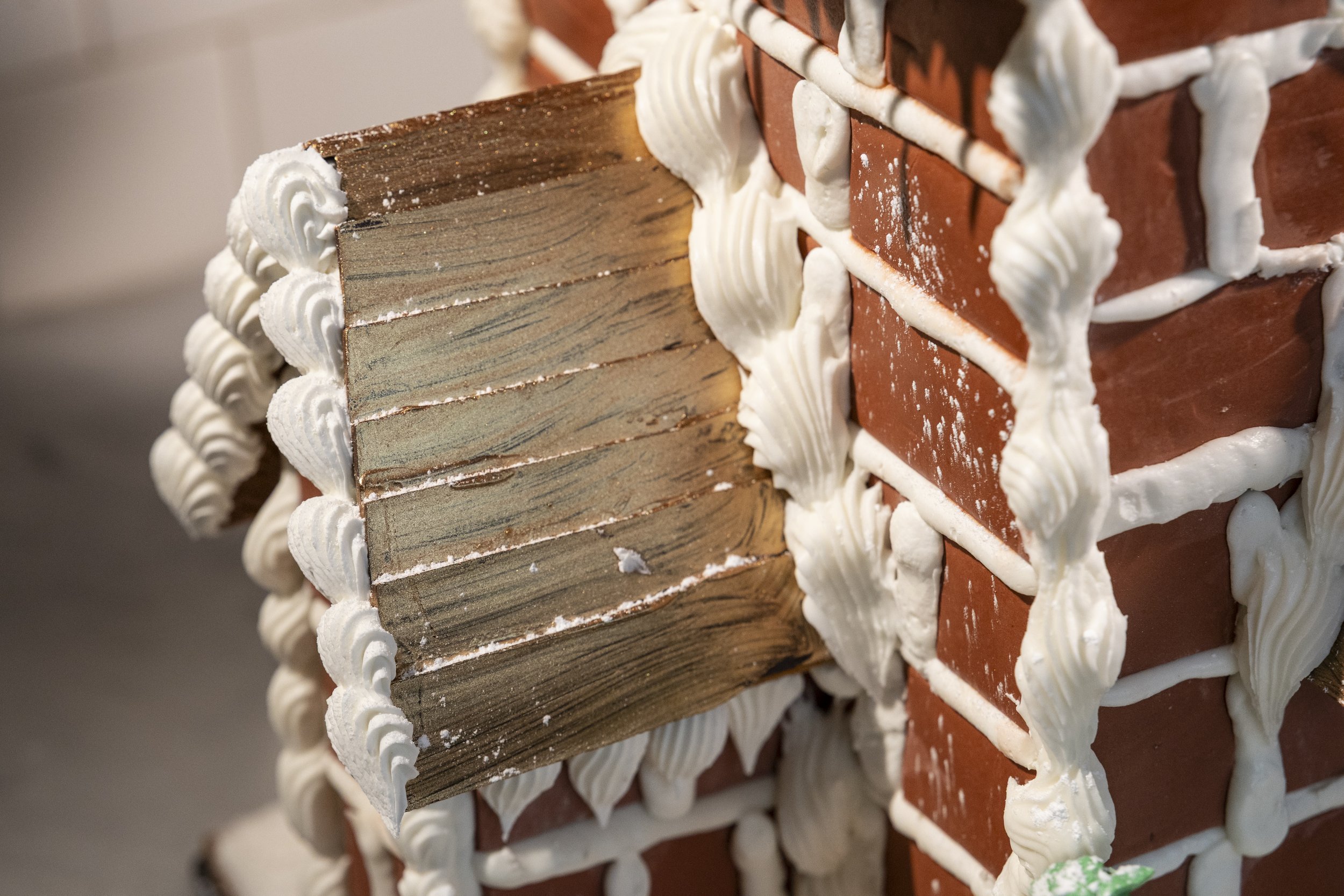 Close-up of the side of a gingerbread house with white icing and red bricks, showing the wooden support structure.
