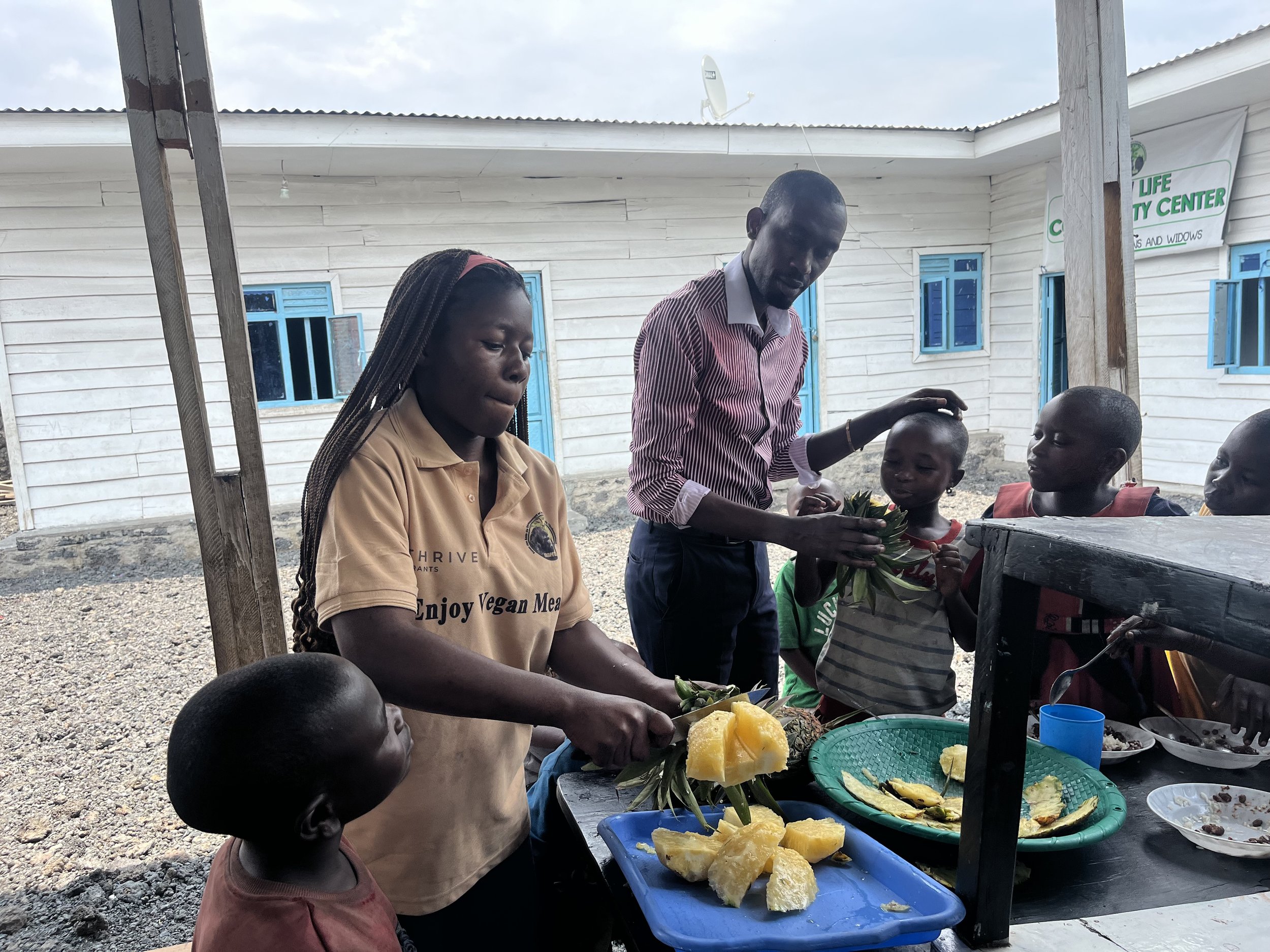A group of children and adults gather around a food stand outdoors, with two trays of pineapple slices and people selecting fruit, in front of a building with blue windows and a sign reading 'Life Outreach Center'.