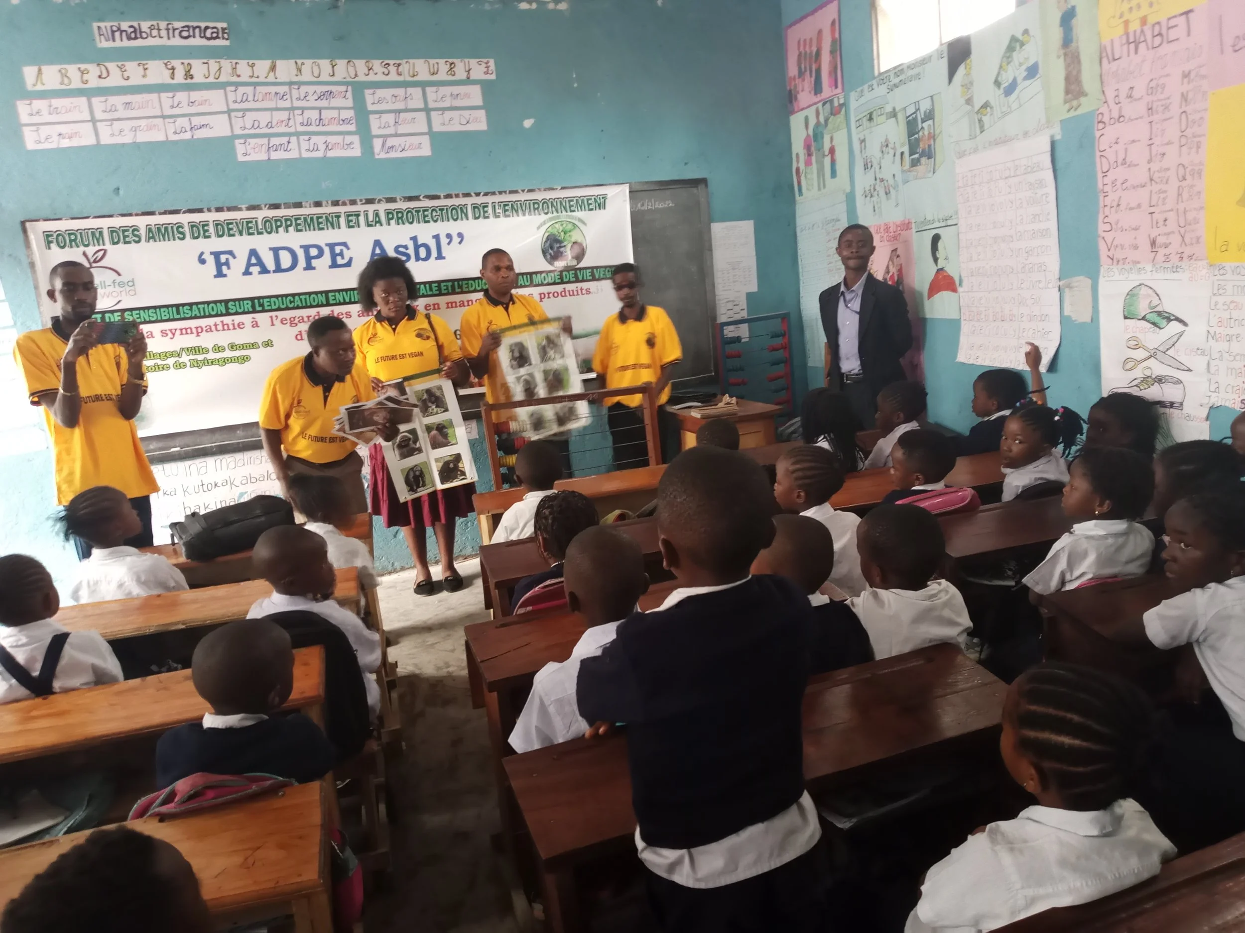 Students in a classroom listening to a presentation about environmental protection, with teachers wearing yellow shirts and colorful posters and handwritten notes on the walls.