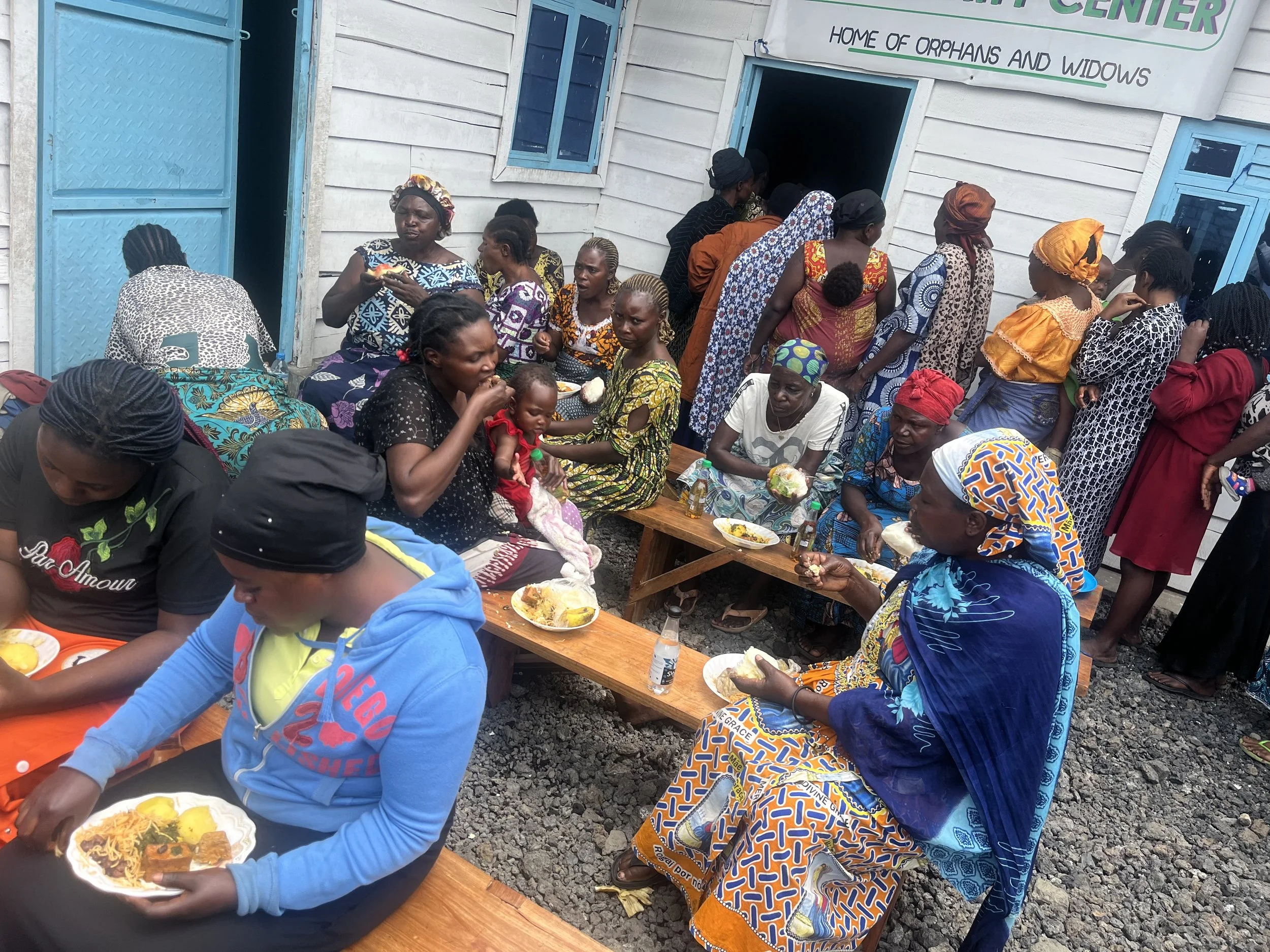 Group of women and children gathering outside a building with a sign that reads 'Home of Orphans and Widows,' enjoying a meal together.