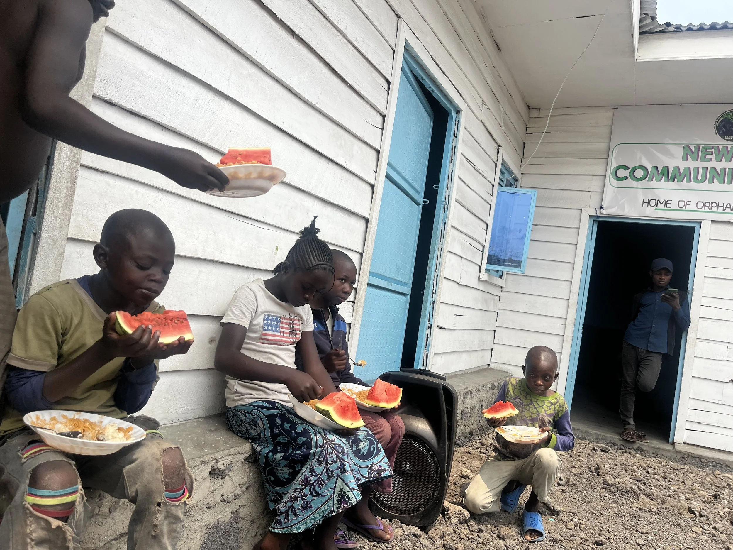 Group of children sitting and standing outside a wooden building, eating watermelon and food, with an adult woman in the background holding a plate of watermelon and a person inside the building looking out at them.