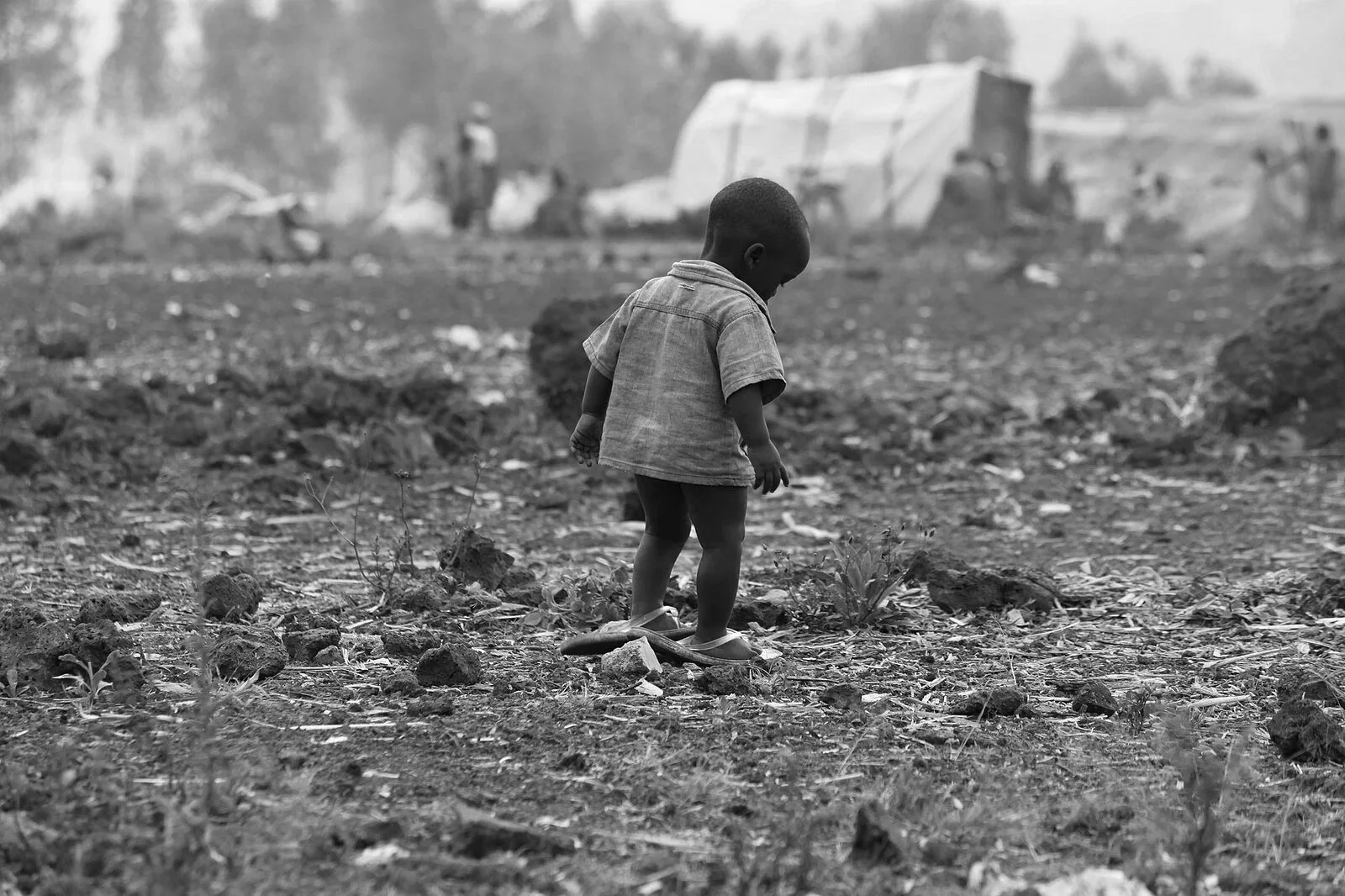 A young child with a short haircut, wearing a shirt, shorts, and flip-flops, standing on uneven ground with rocks and sparse plants in a desolate landscape.