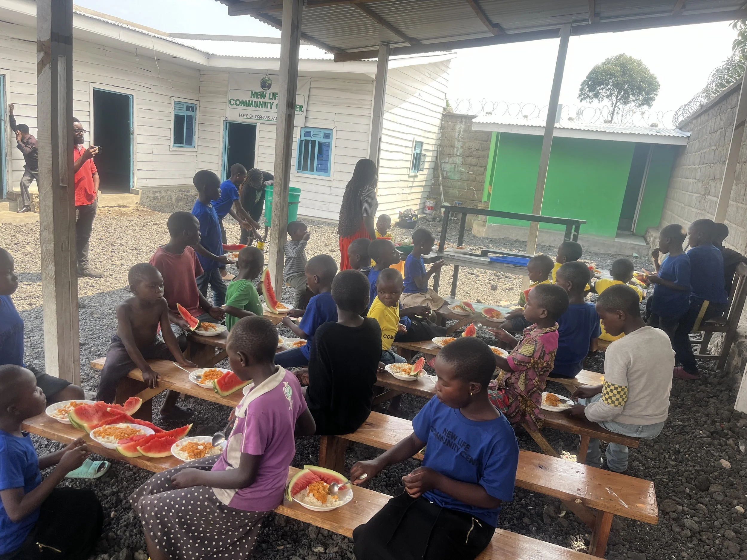 Children sitting on wooden benches having a meal, with plates of food and slices of watermelon, under a makeshift shelter outside a community center.
