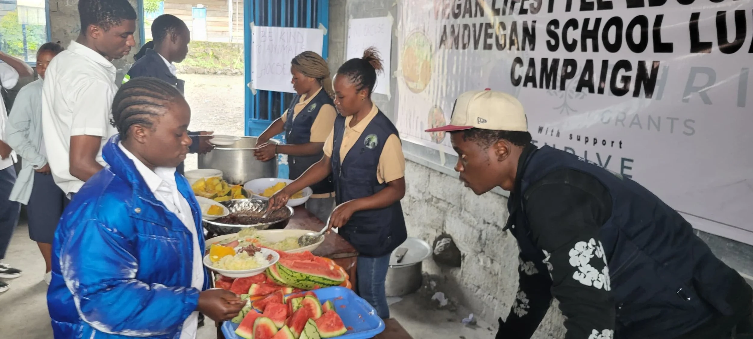 Students and volunteers serving food at a school lunch event, with watermelons and other dishes on a table, and a large banner in the background promoting a vegan school lunch campaign.