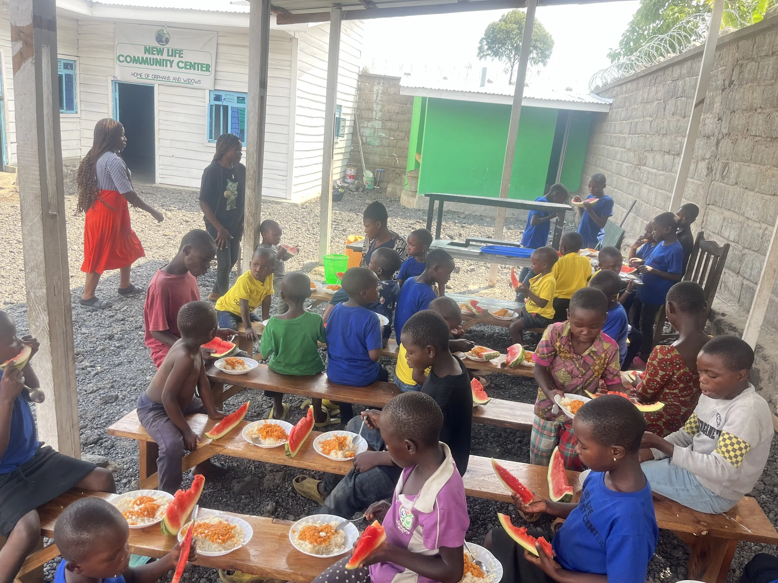 Children sitting outdoors on wooden benches eating watermelon, with plates of rice and vegetables, at a community center in a rural area, featuring a sign for New Life Community Center in the background.