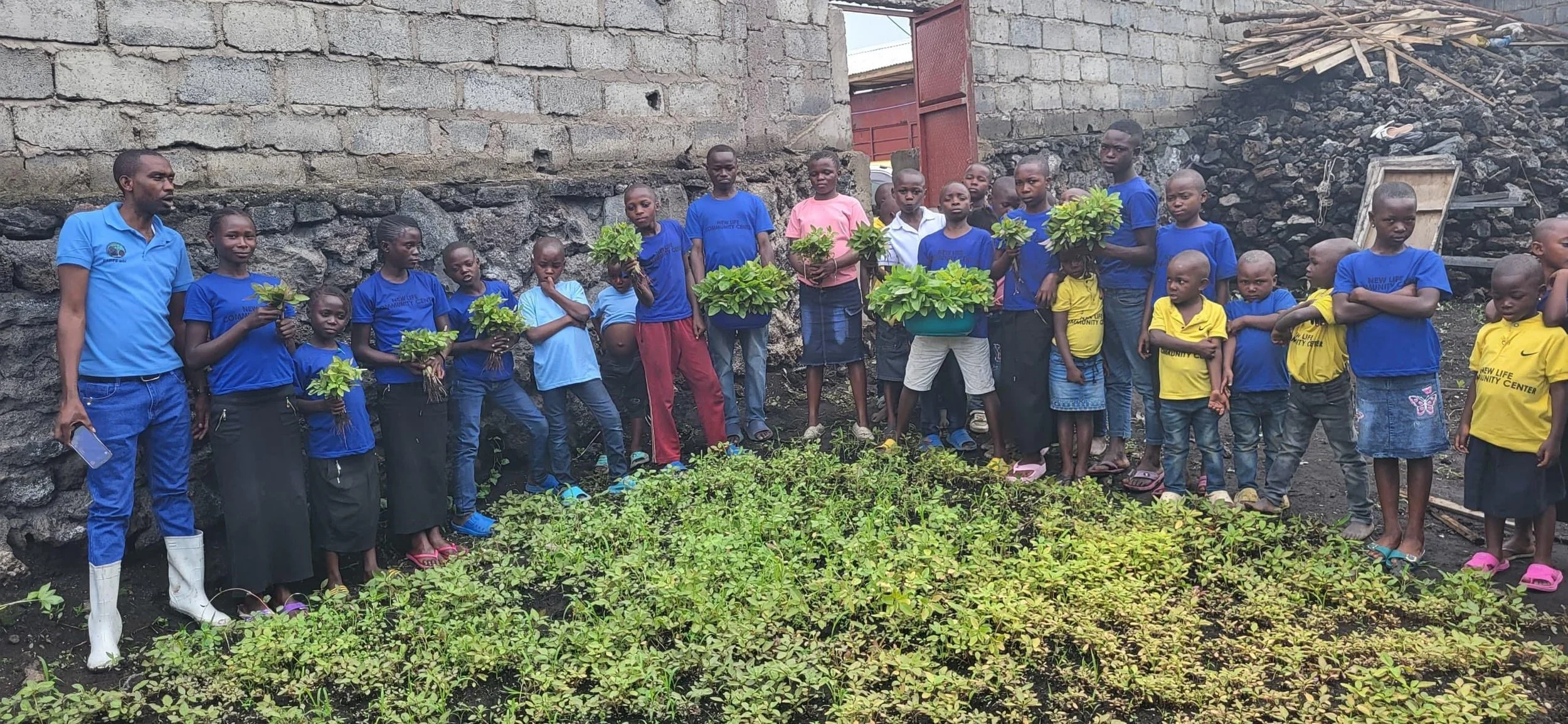 Group of children and young adults standing in a row in an outdoor garden, some holding plants, with a stone wall and pile of wood in the background.