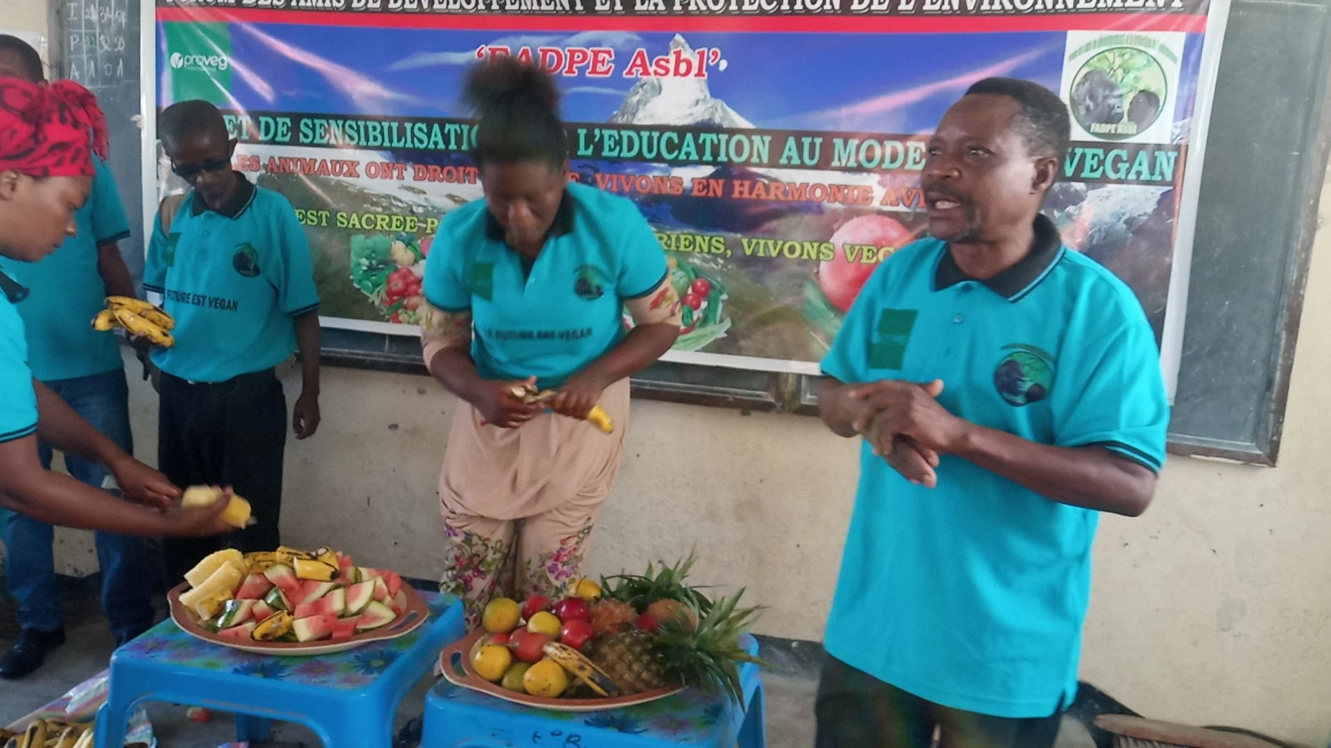 A group of people participating in a fruit-themed event or presentation, with fruits like bananas, watermelons, pineapples, and apples displayed on tables. One person is holding bananas, and there is a large colorful banner in the background with text in French.