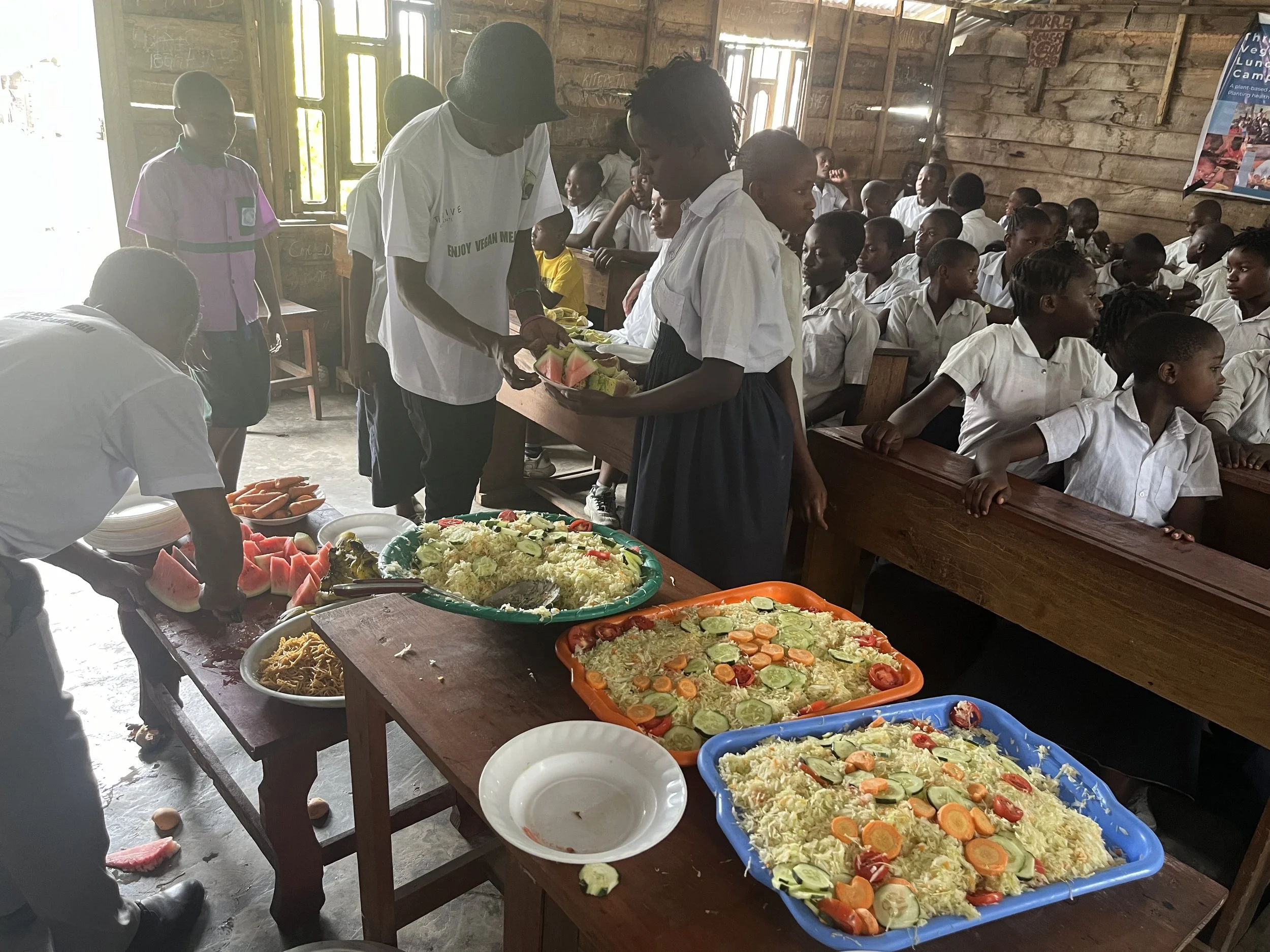 A group of students in white uniforms seated in desks inside a classroom, with a table of food including rice, vegetables, and fruit in front of them, and some people serving or preparing food near the table.