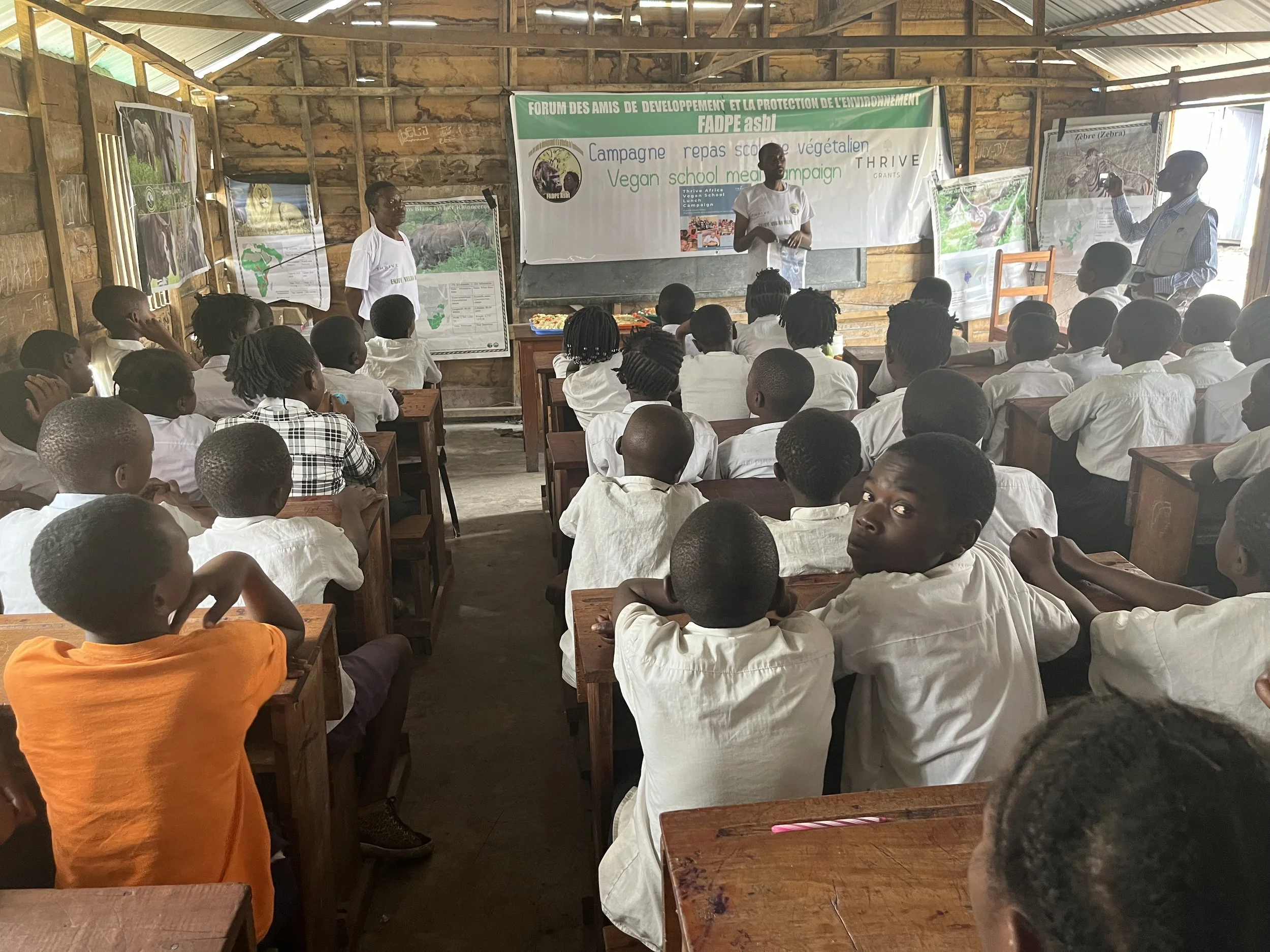 A classroom with young students seated at wooden desks, listening to a presentation by teachers at the front. The classroom is made of wood with educational posters on the walls. A banner is visible behind the teachers.