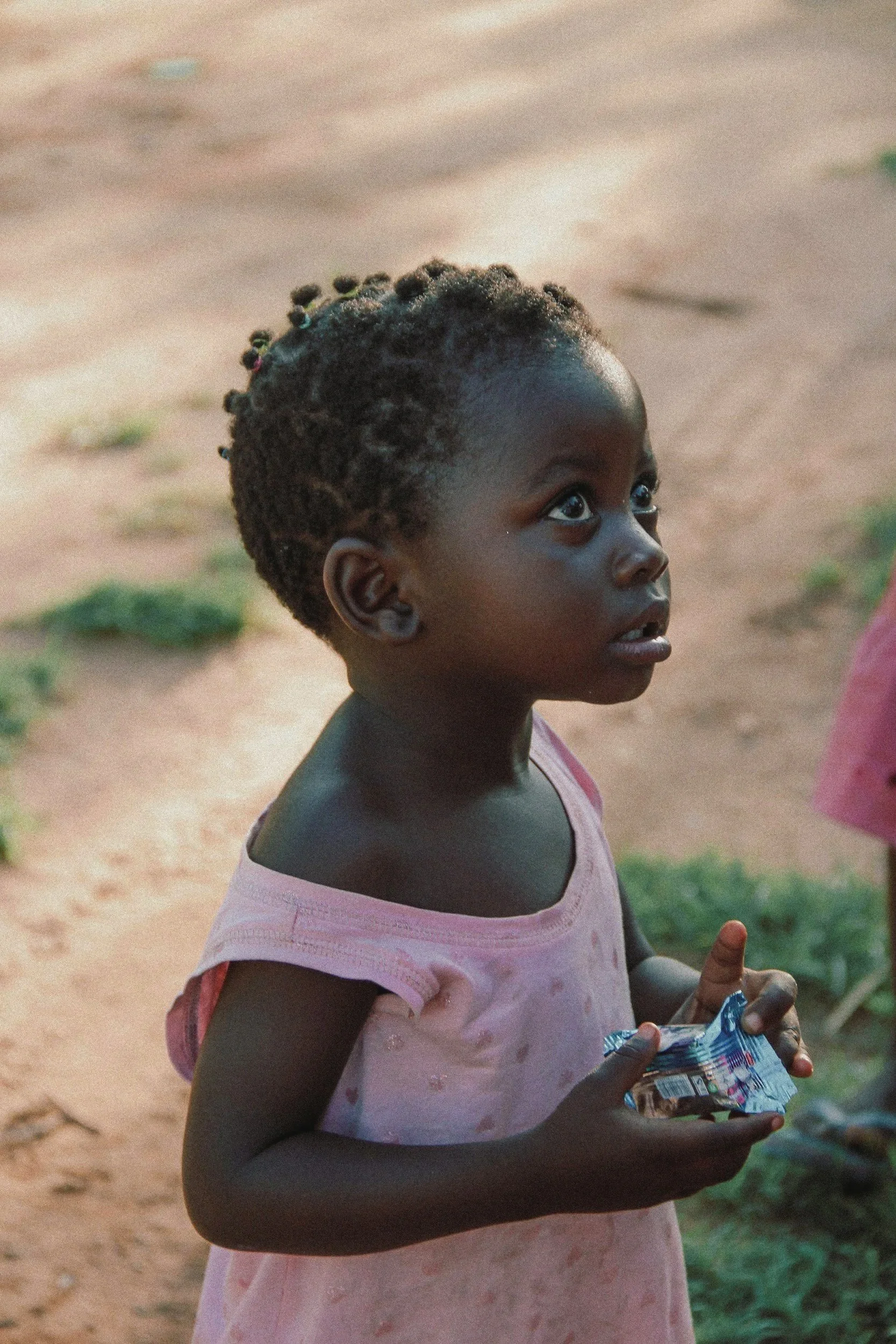 A young girl with dark skin and curly hair styled with small beads, wearing a pink dress, looking upward with a curious expression while holding a small snack or candy wrapper in her hand.