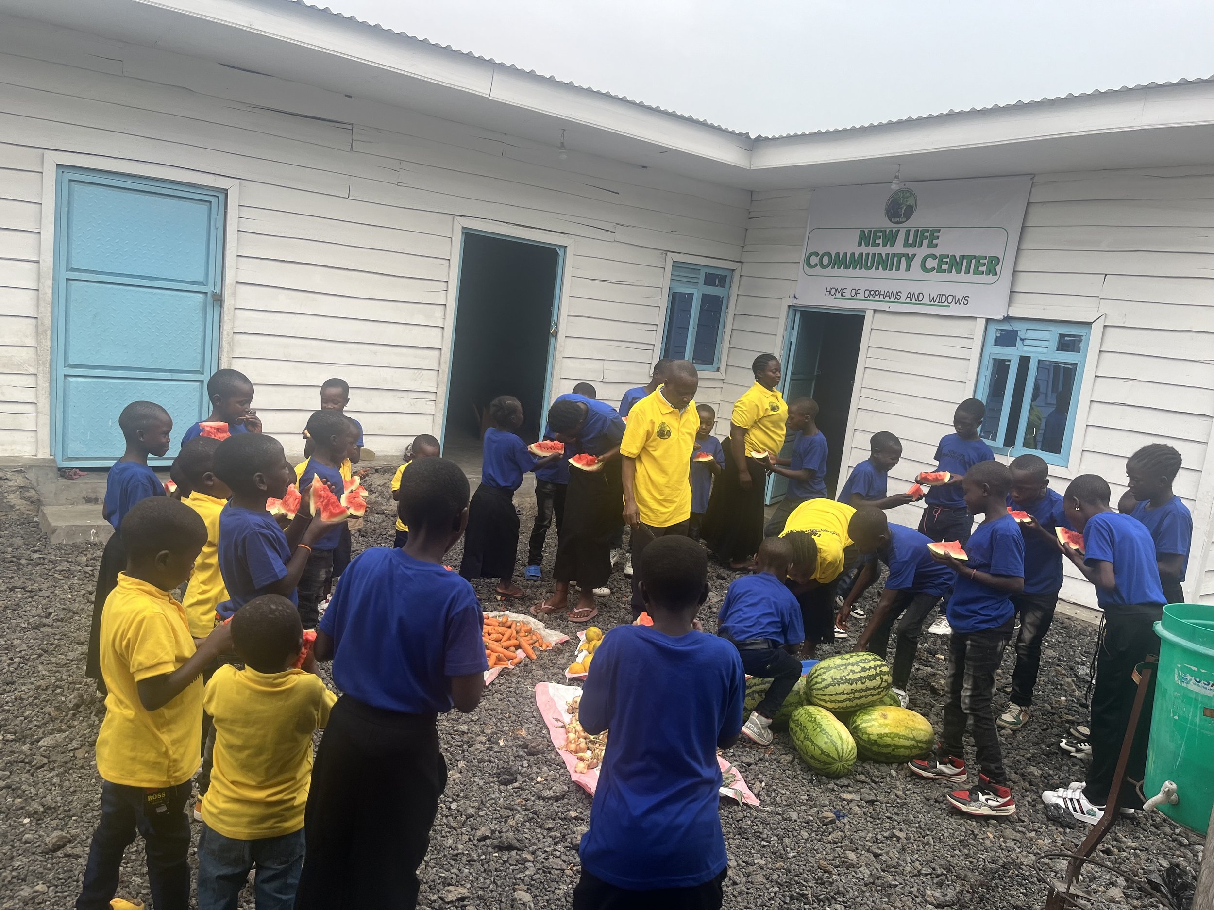 Children and adults gathered outside the New Life Community Center, holding slices of watermelon and surrounded by watermelons, carrots, and other vegetables, during a communal meal or event.