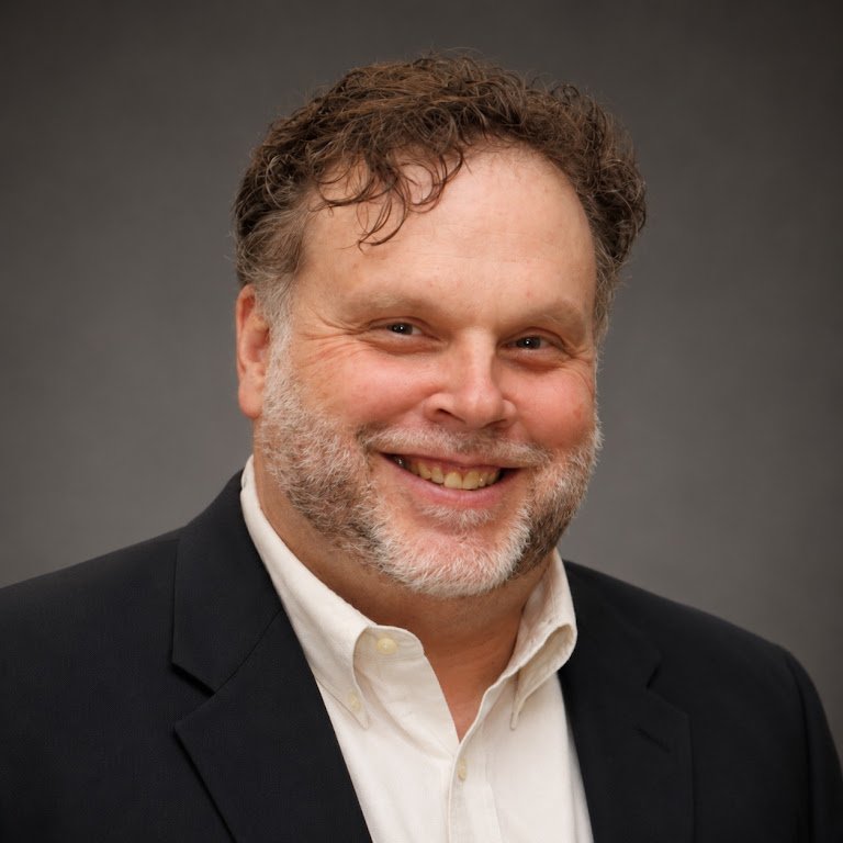 A smiling man with curly hair and a beard in a dark suit and white shirt
