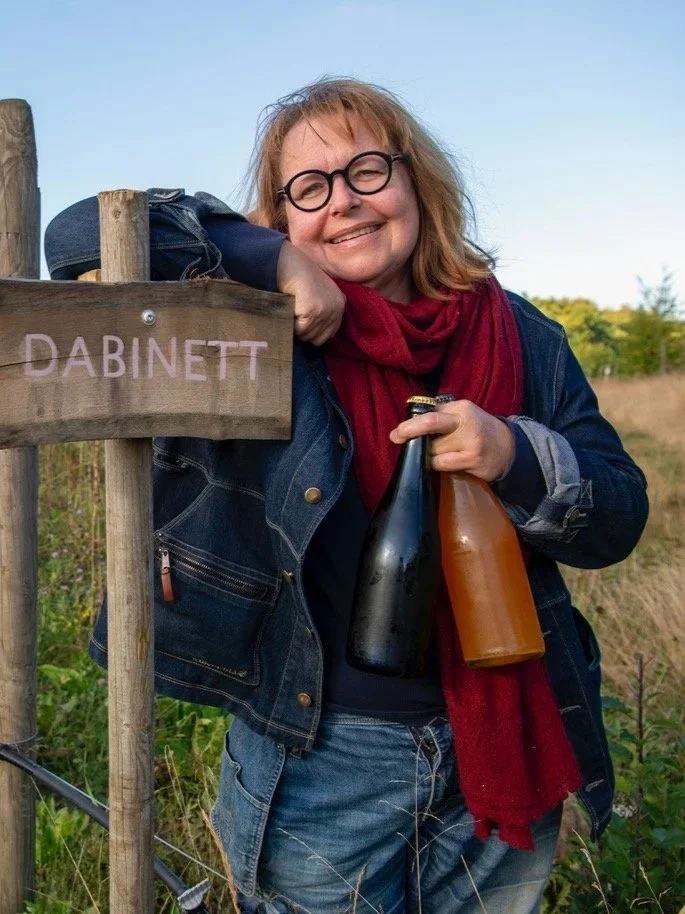 Kvinna leende med solglasögon, röd scarf, jeansjacka, håller två flaskor, står bredvid en skylt med texten DABINN. Utomhus, natur i bakgrunden.