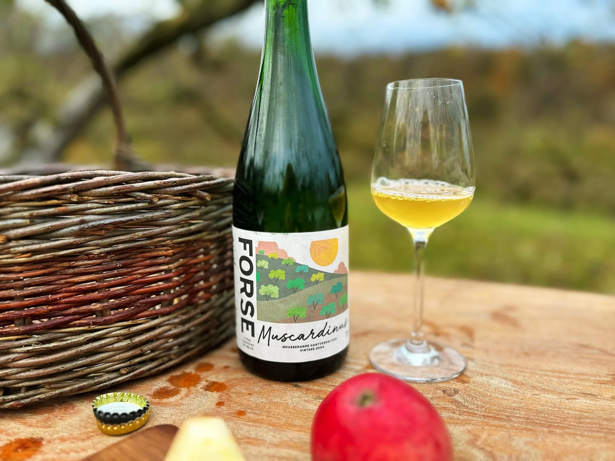 En vinflaska med etiketten 'Forsse Muscadin' och ett glas med gul dryck på ett träbord utomhus, en röd äpple och en grillad bananskiva i förgrunden, en gallerkorg till vänster.