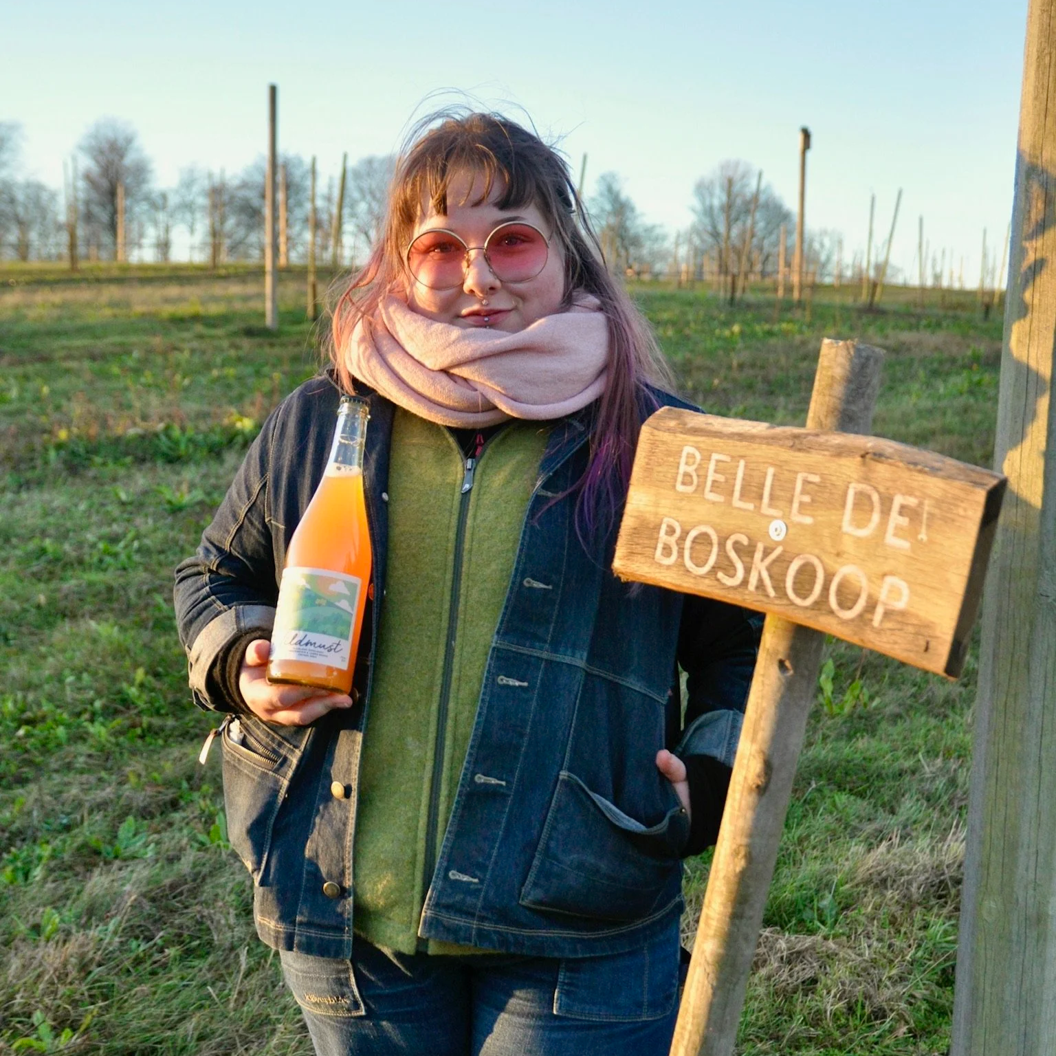 En kvinna med rosa glasögon och en scarf runt halsen står utomhus i ett grönt område, håller i en flaska med läsk och står bredvid en träskylt som säger 'Belle del boskoop'.