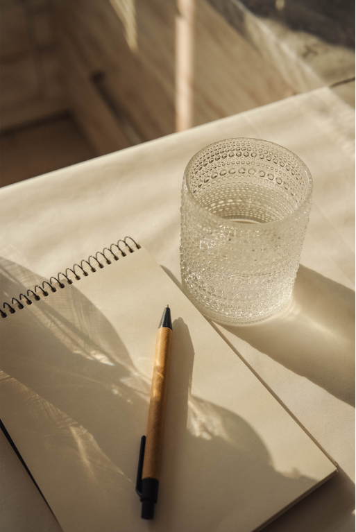 A spiral-bound notebook with blank pages, a wooden pen with a black tip, and a textured transparent glass of water on a light-colored surface, with sunlight casting shadows.