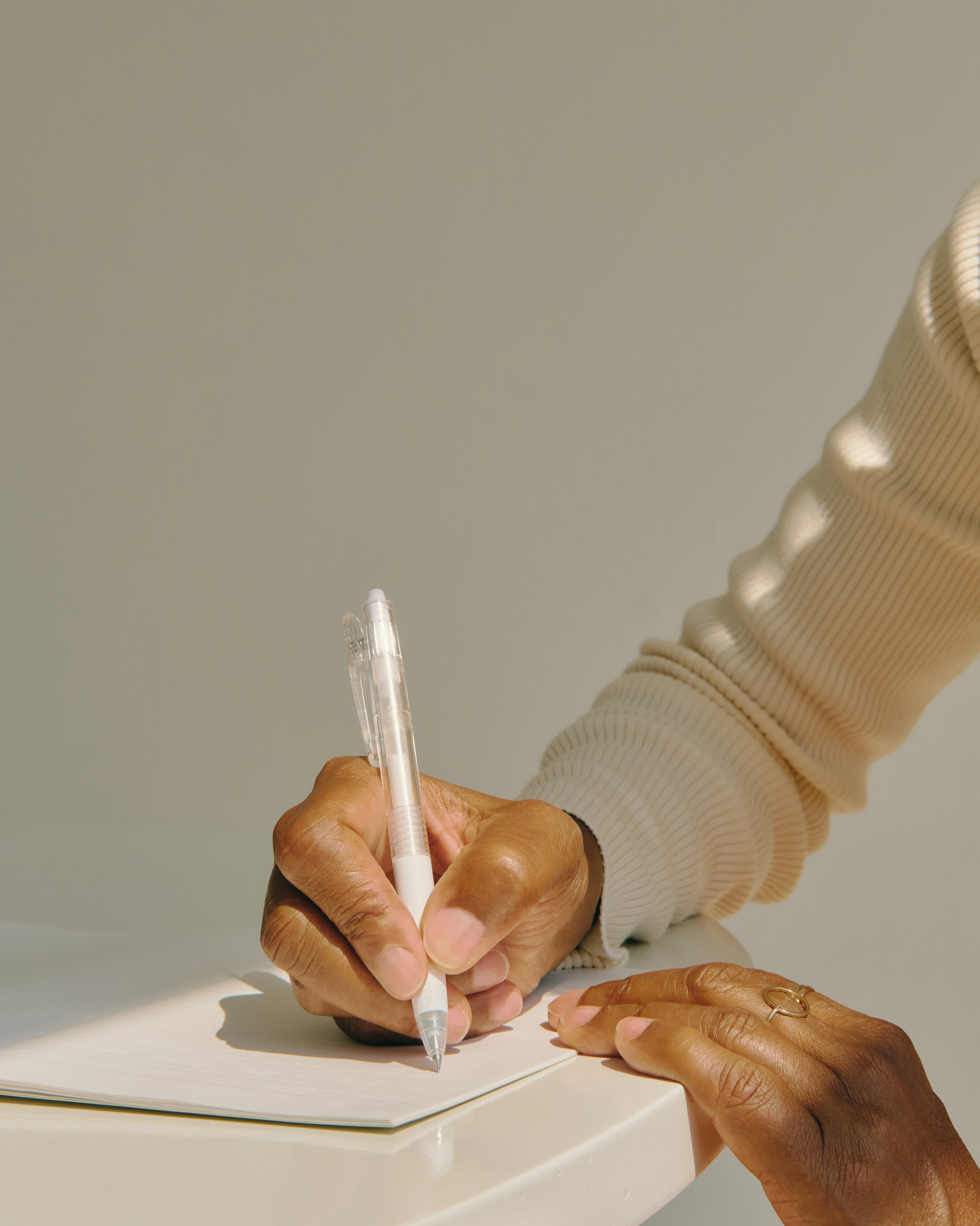 Person writing in a notebook with a pen, wearing a beige ribbed sweater, with natural light illuminating the scene.