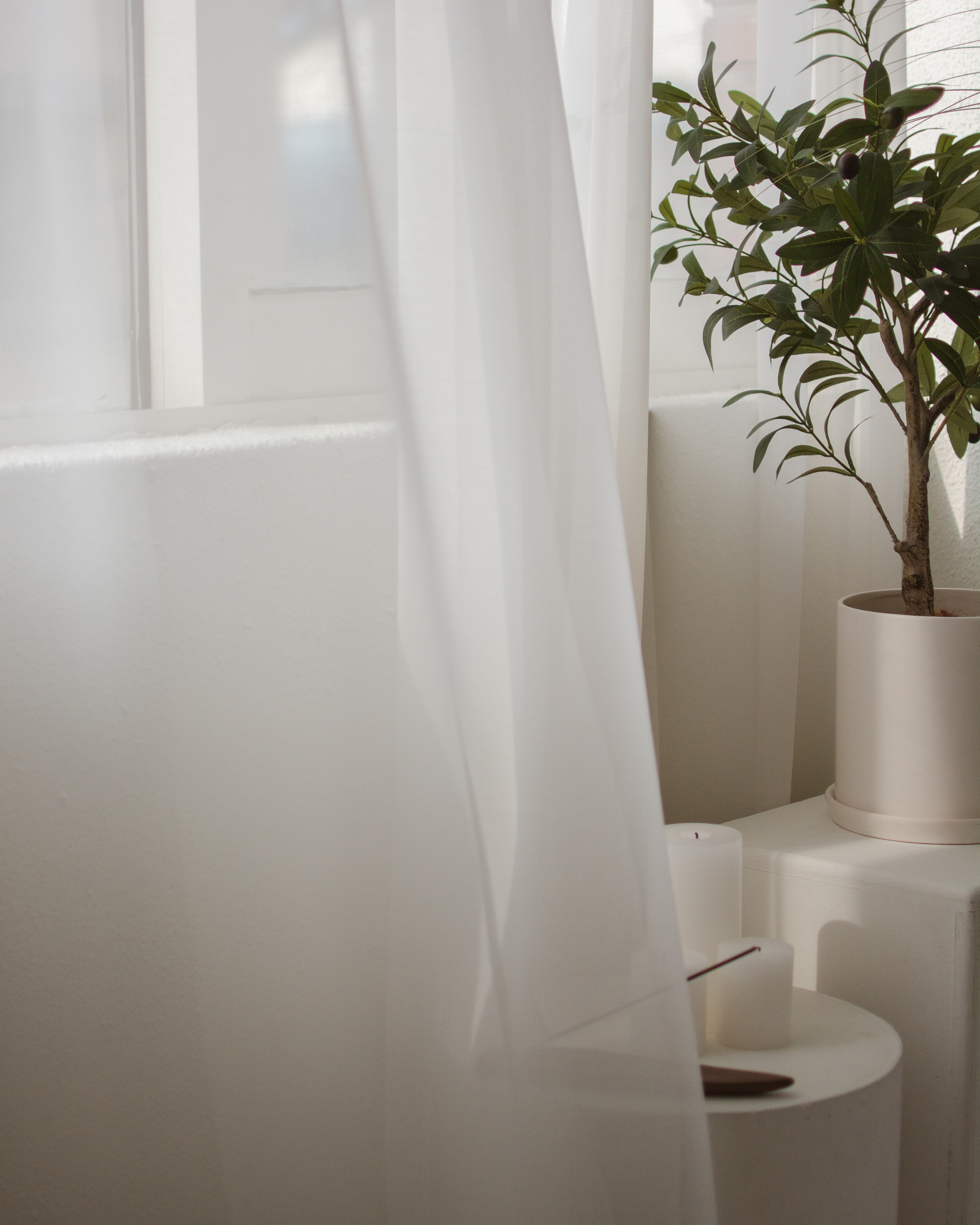 Soft white curtains partially obscuring a potted green plant on a white surface with candles and a stick on a white table in a bright, minimalistic room.