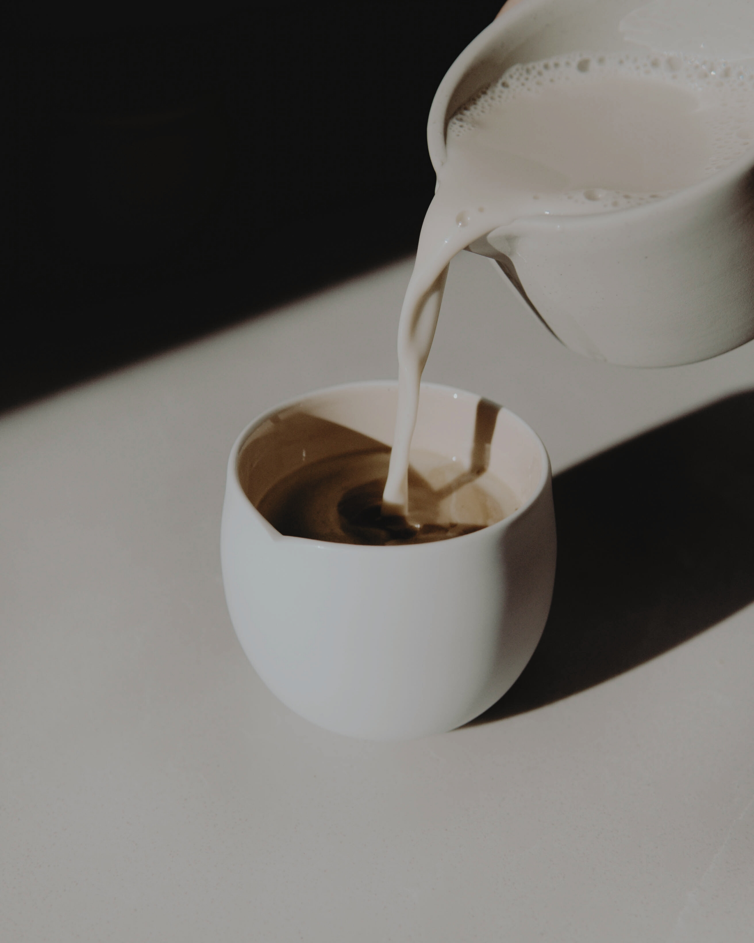 Creamy milk being poured from a pitcher into a white cup.