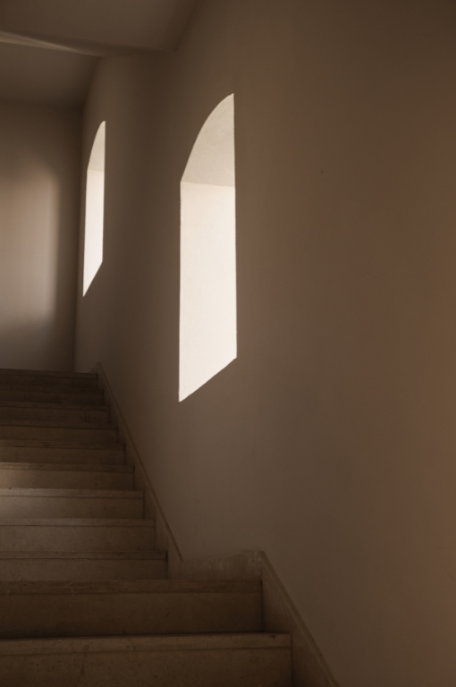 Sunlight cast through arched windows creating shadows on a white interior wall near a staircase.