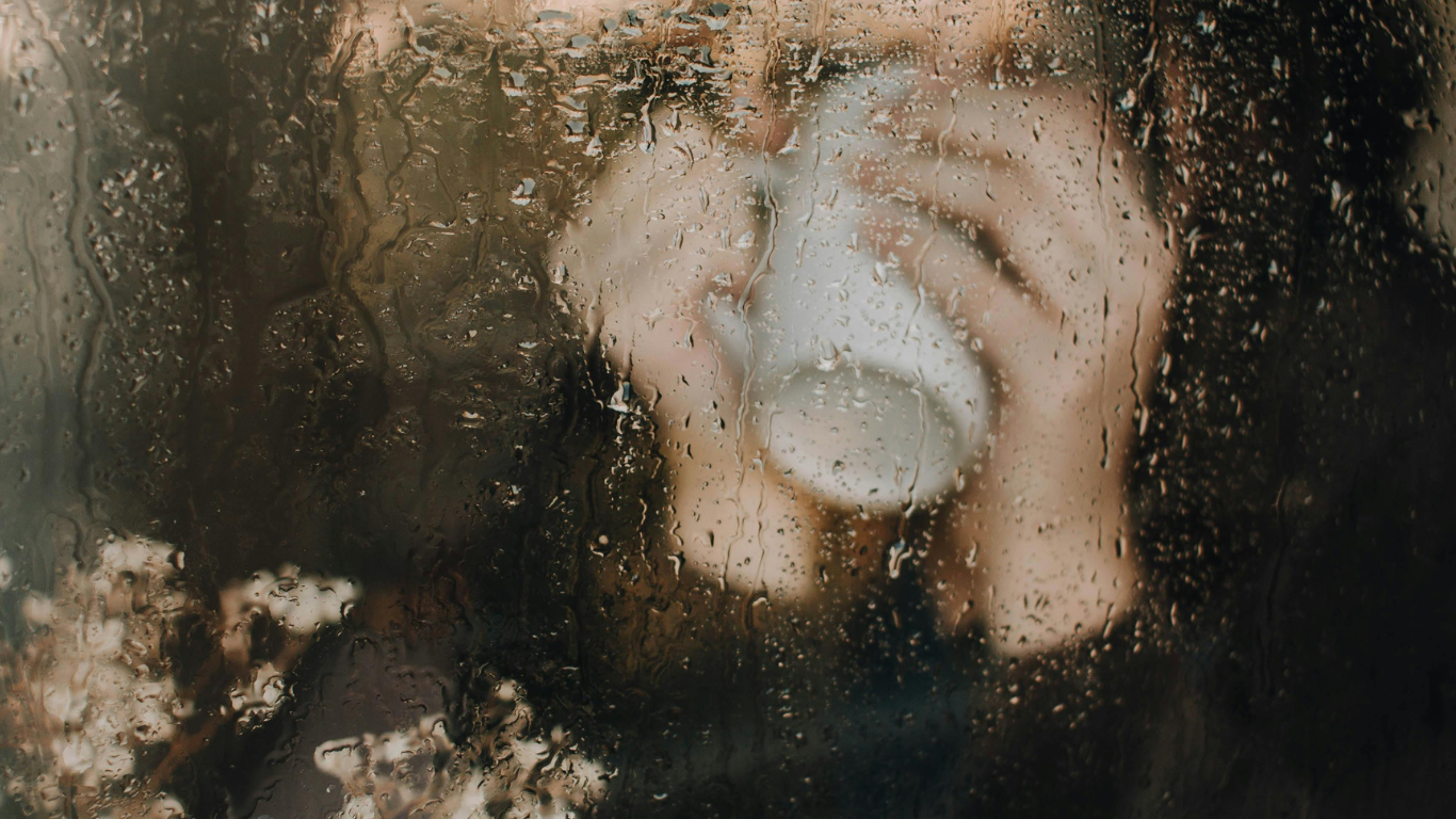 Person with curly hair wearing a face mask, seen through a rain-covered window, holding a cup.