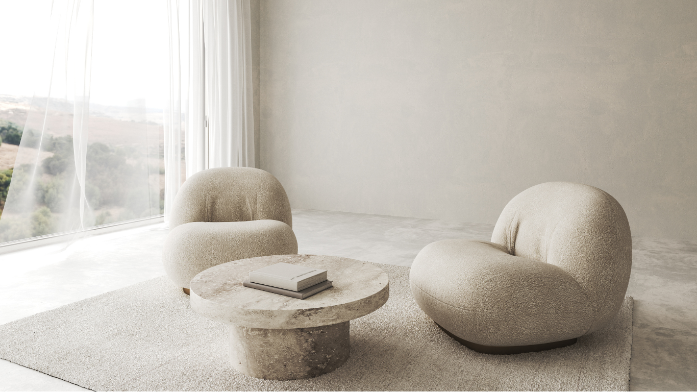 Minimalist living room with two rounded beige armchairs, a round stone coffee table with books, light-colored walls, and large windows with sheer curtains letting in natural light.
