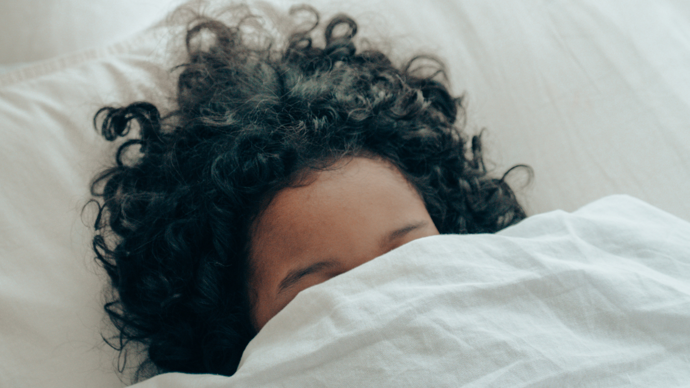 A person with curly hair sleeping under a white blanket.