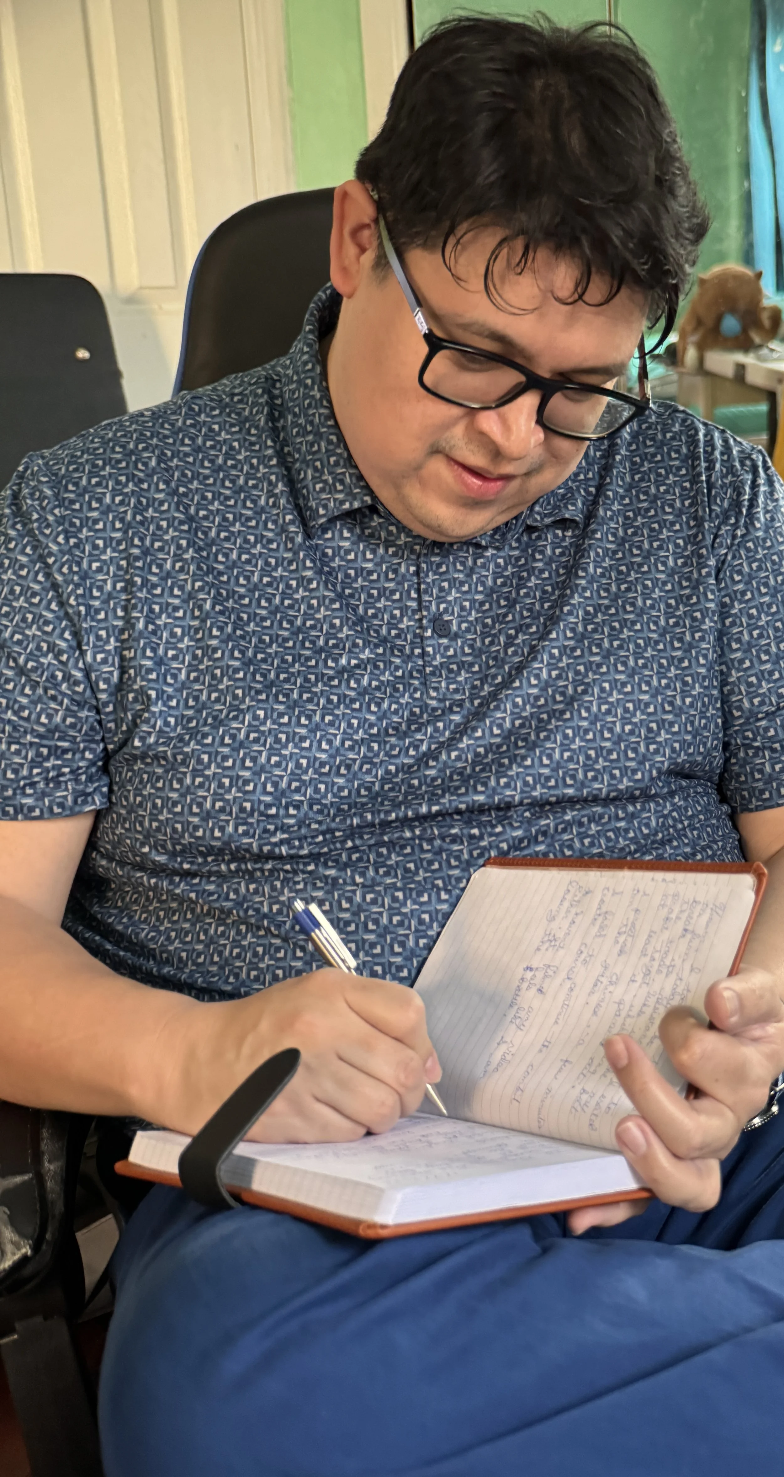 A man with glasses, wearing a patterned blue shirt, sitting on a chair, writing in a notebook with a blue and silver pen, in a room with green walls and some objects in the background.