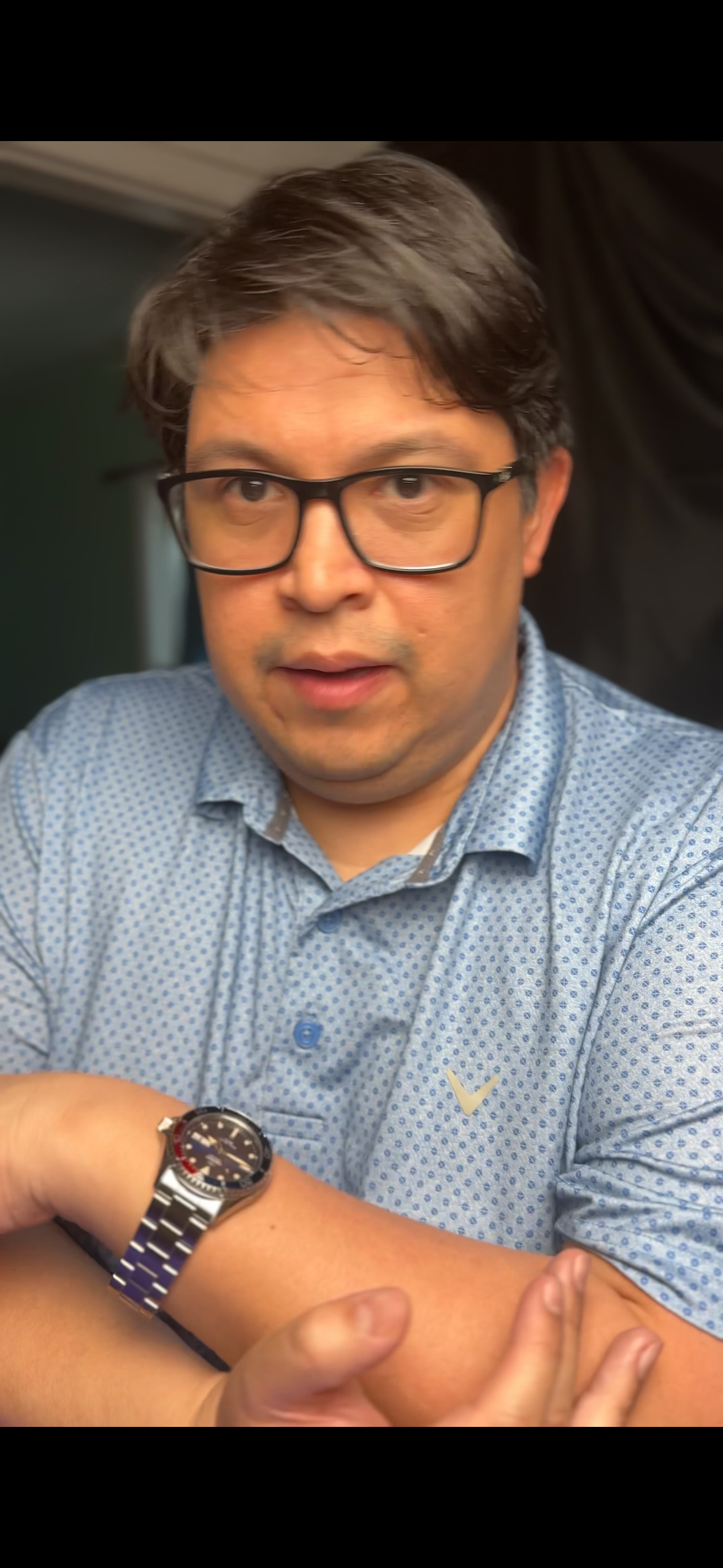 A man wearing glasses and a light blue patterned shirt with a gold check mark logo, adjusting his watch on his left wrist, looking into the camera.