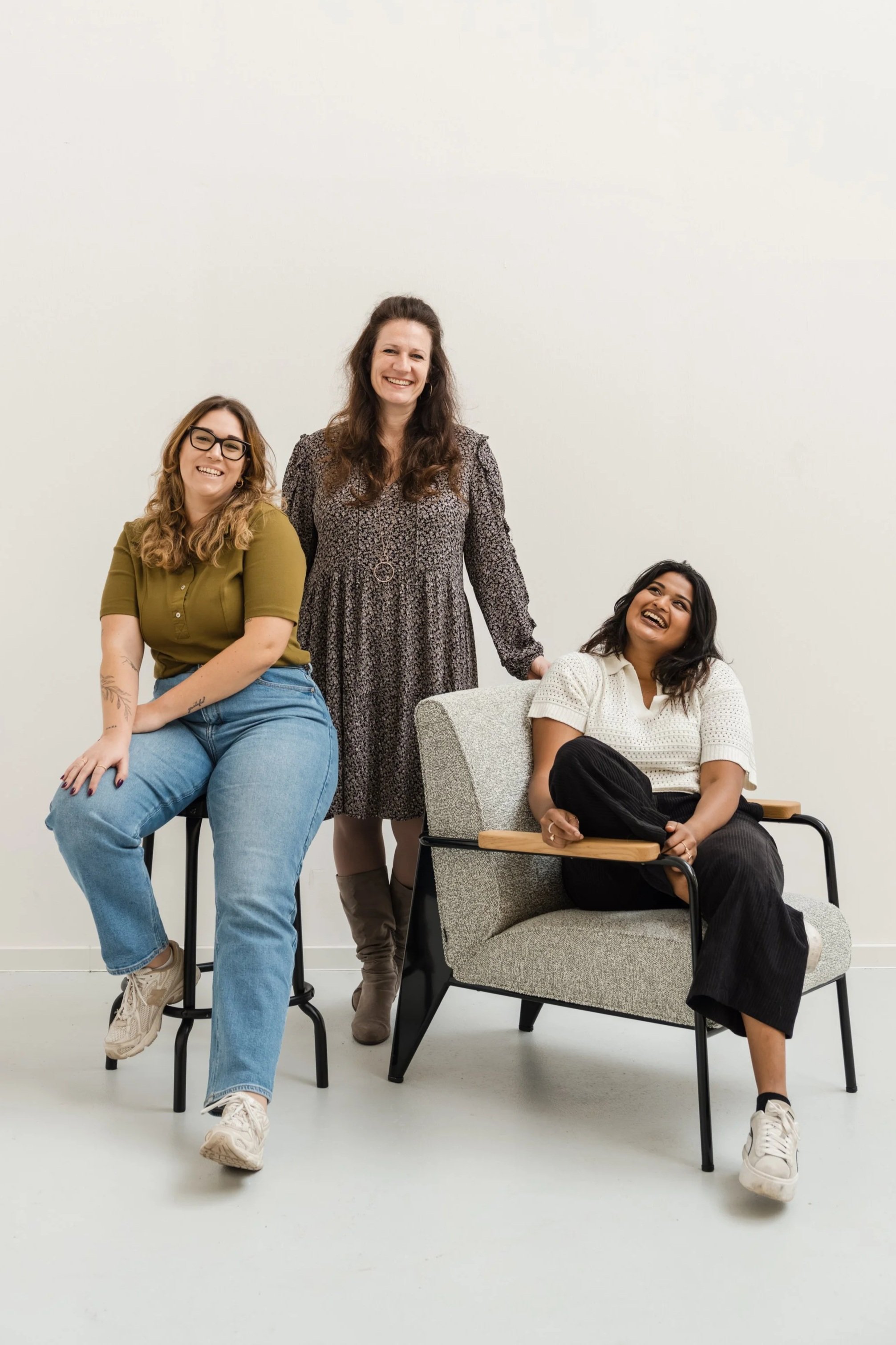 Drei lachende Frauen in einem Raum, eine sitzt auf einem Stuhl, eine steht und die andere sitzt in einem Sessel, alle wirken entspannt und fröhlich.