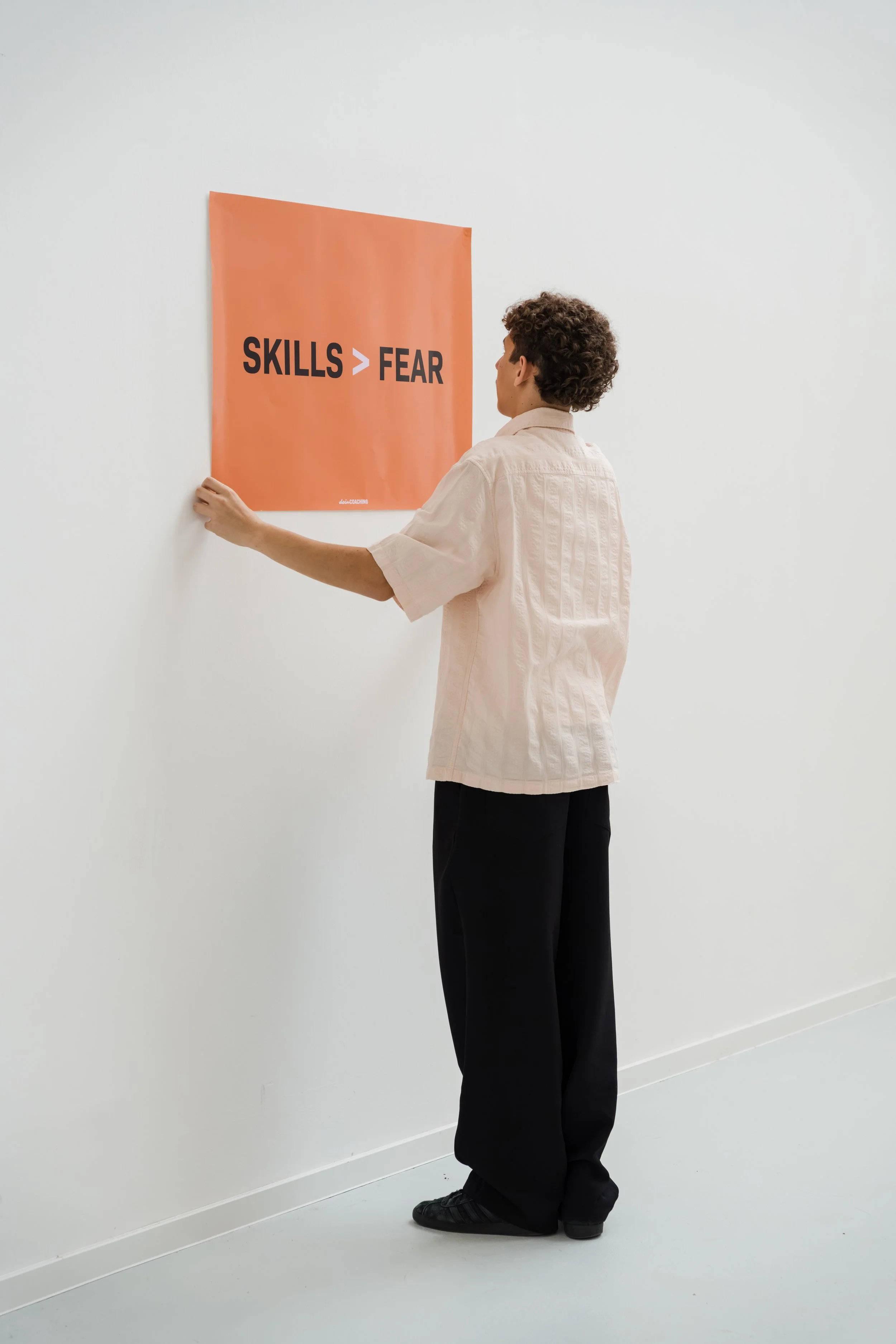 Junge Person hängt ein Poster mit der Aufschrift "Skills > Fear" an die weiße Wand in einem modernen Raum.