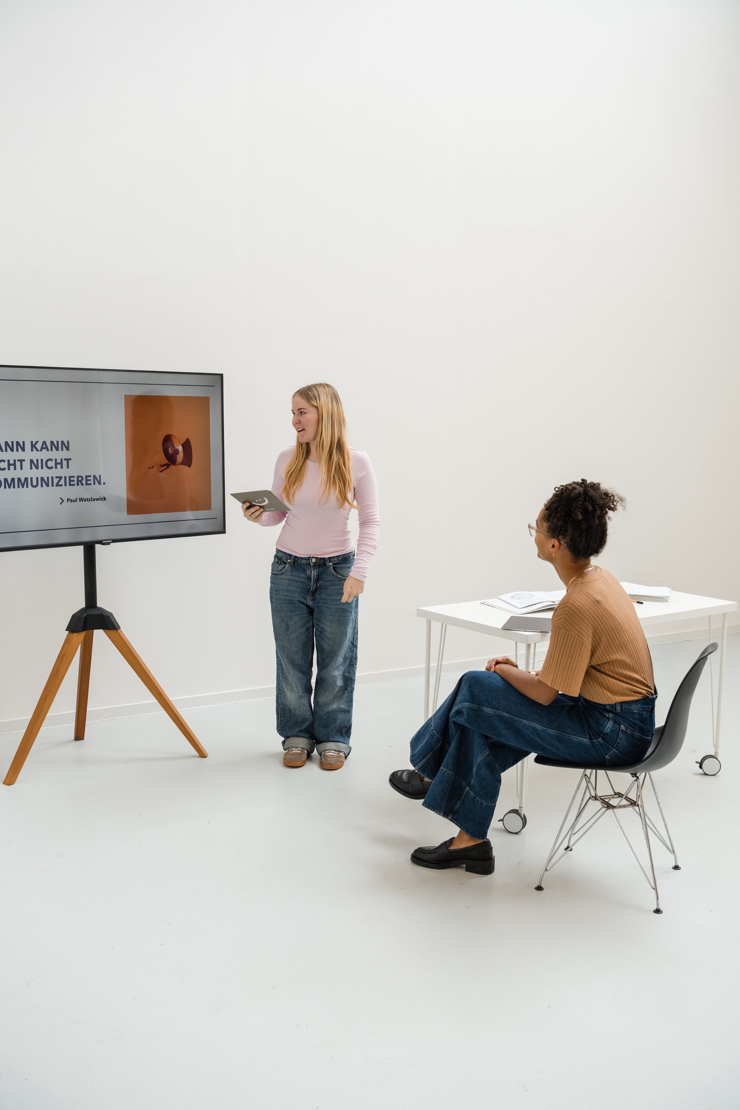 Zwei Frauen in einem weißen Raum, eine präsentiert vor einem Bildschirm, die andere sitzt an einem Tisch und hört zu.