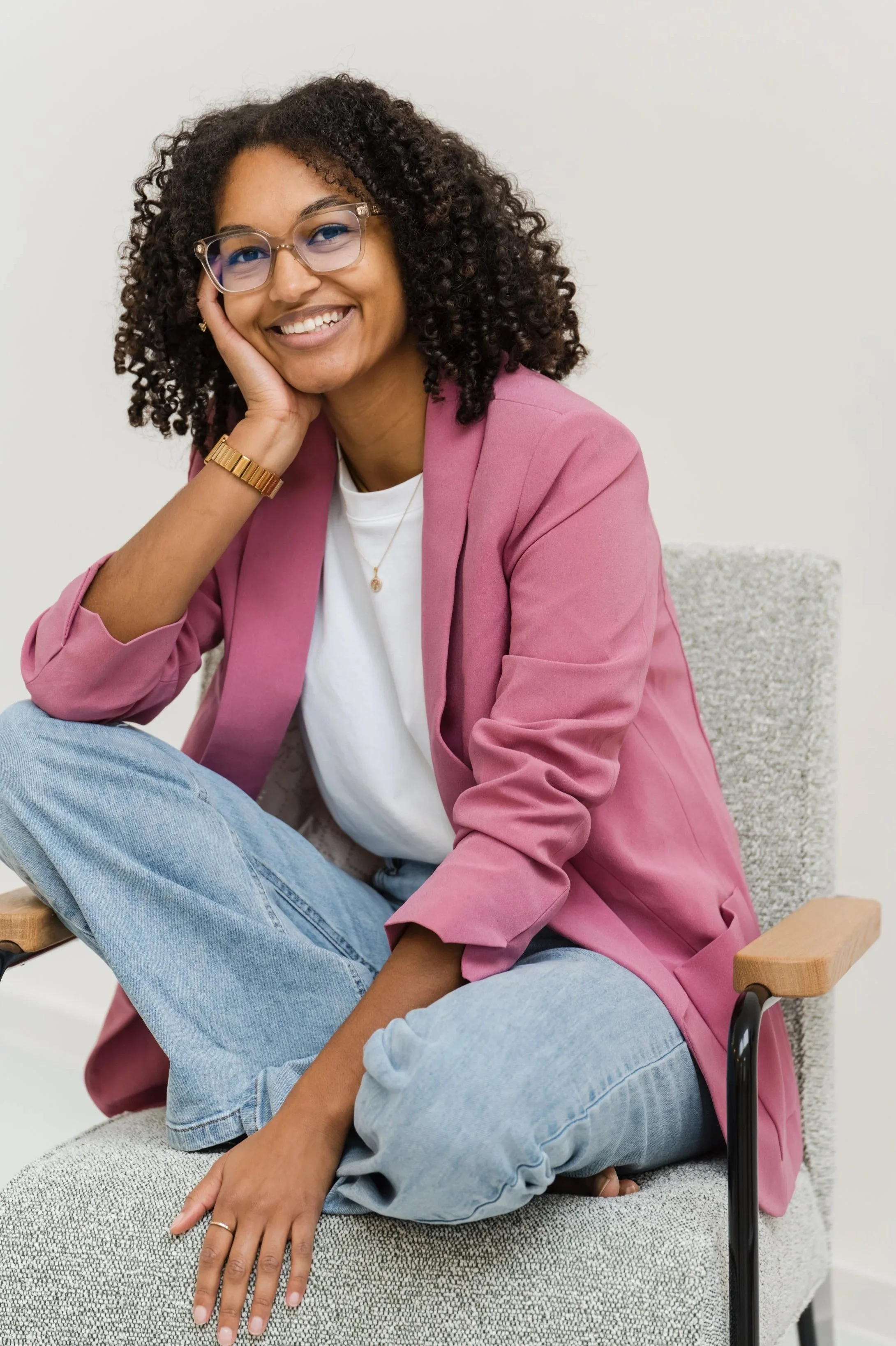 Junge Frau mit Brille, lächelnd, trägt pinken Blazer und Jeans, sitzt auf einem Stuhl, hat lockige Haare, hält den Kopf mit der Hand, in einem hellen Raum.