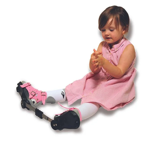 A young girl sitting on the floor, wearing a fixed abduction brace or boots and bar for Ponseti correction of clubfoot.