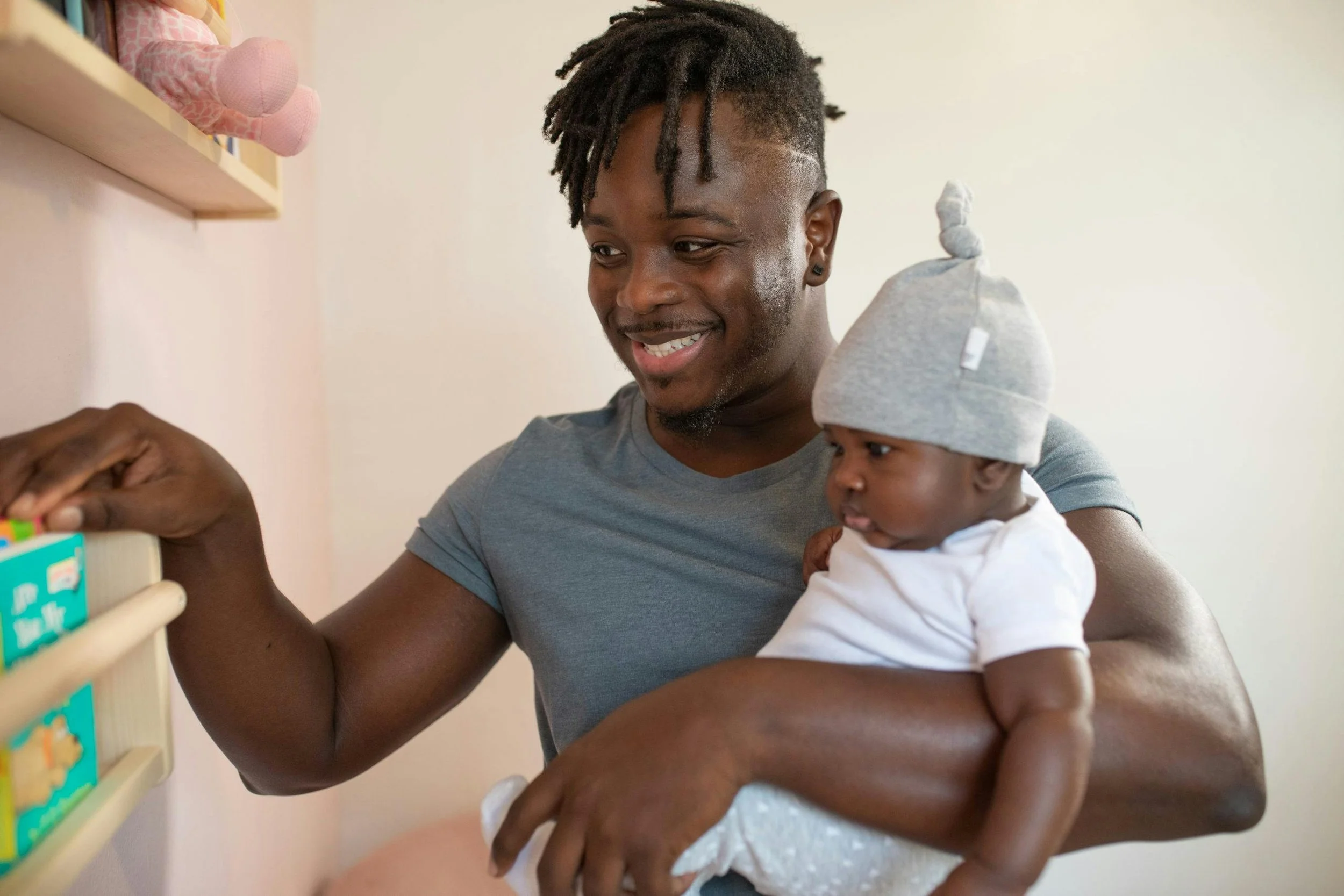A man smiling while holding a baby boy in his arm, both looking at colorful objects on a shelf.