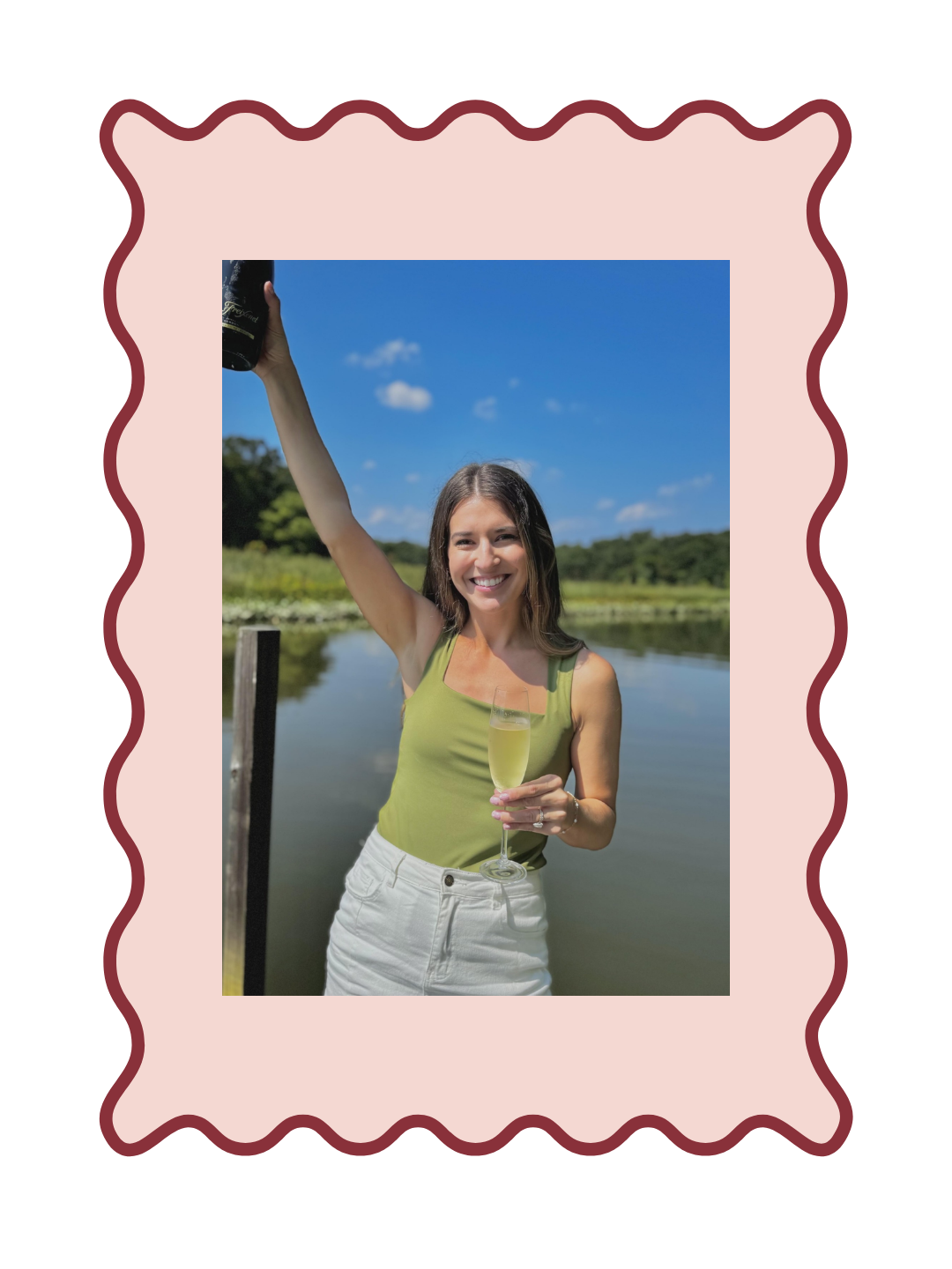 Tierney Robb of Chesapeake Socials holding a glass and bottle of champagne on a dock. The image has a light pink border around it with a dark red scalloped outline to make a frame.