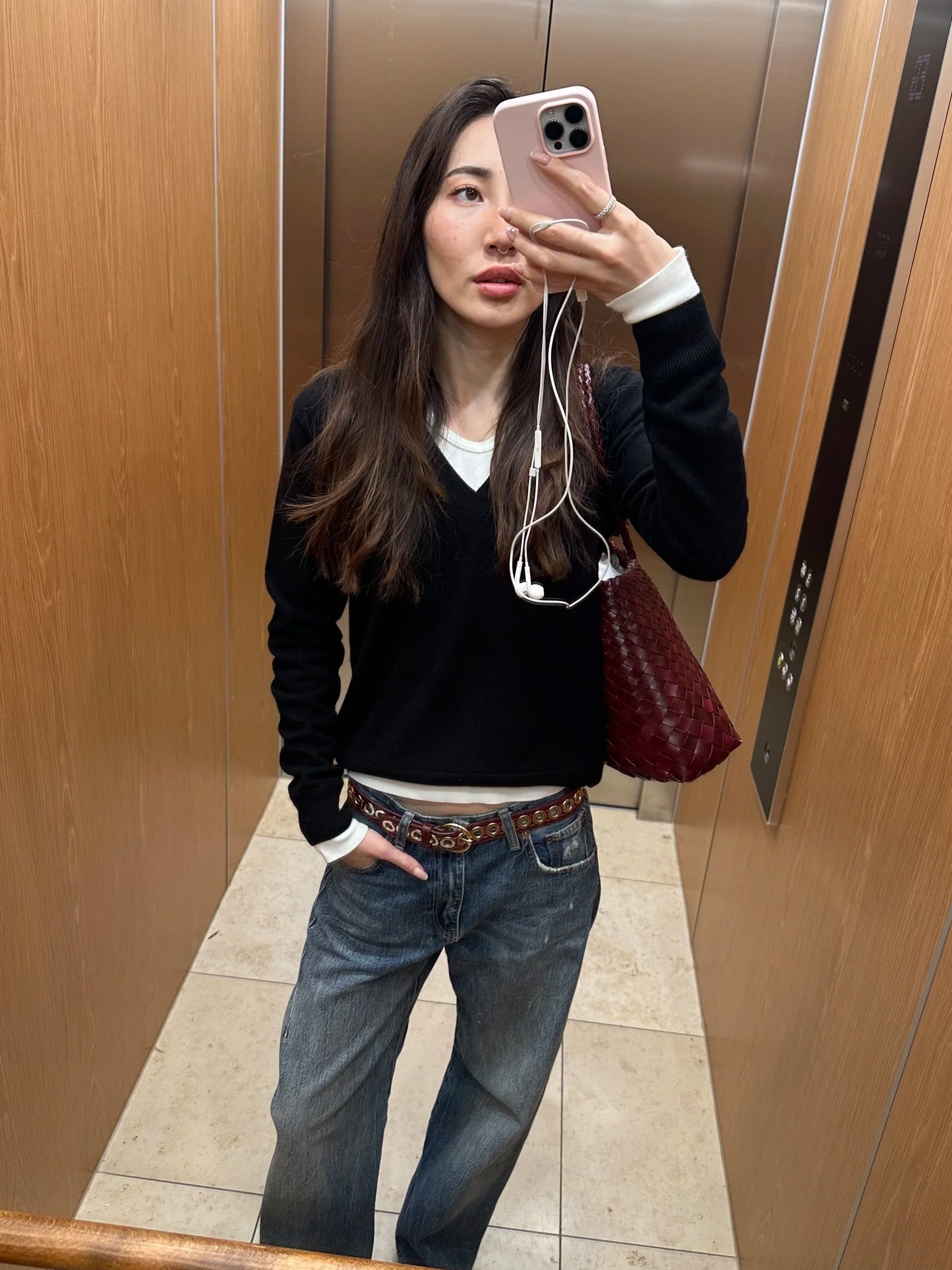 Young woman taking a mirror selfie inside an elevator, holding her phone in front of her face, wearing a black and white sweater, jeans, and carrying a red woven bag.