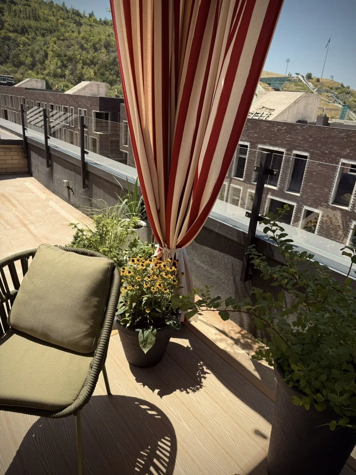 A balcony with a green cushioned chair, potted plants with flowers, and a red-and-white striped curtain, overlooking a cityscape with brick buildings and green hills in the background.