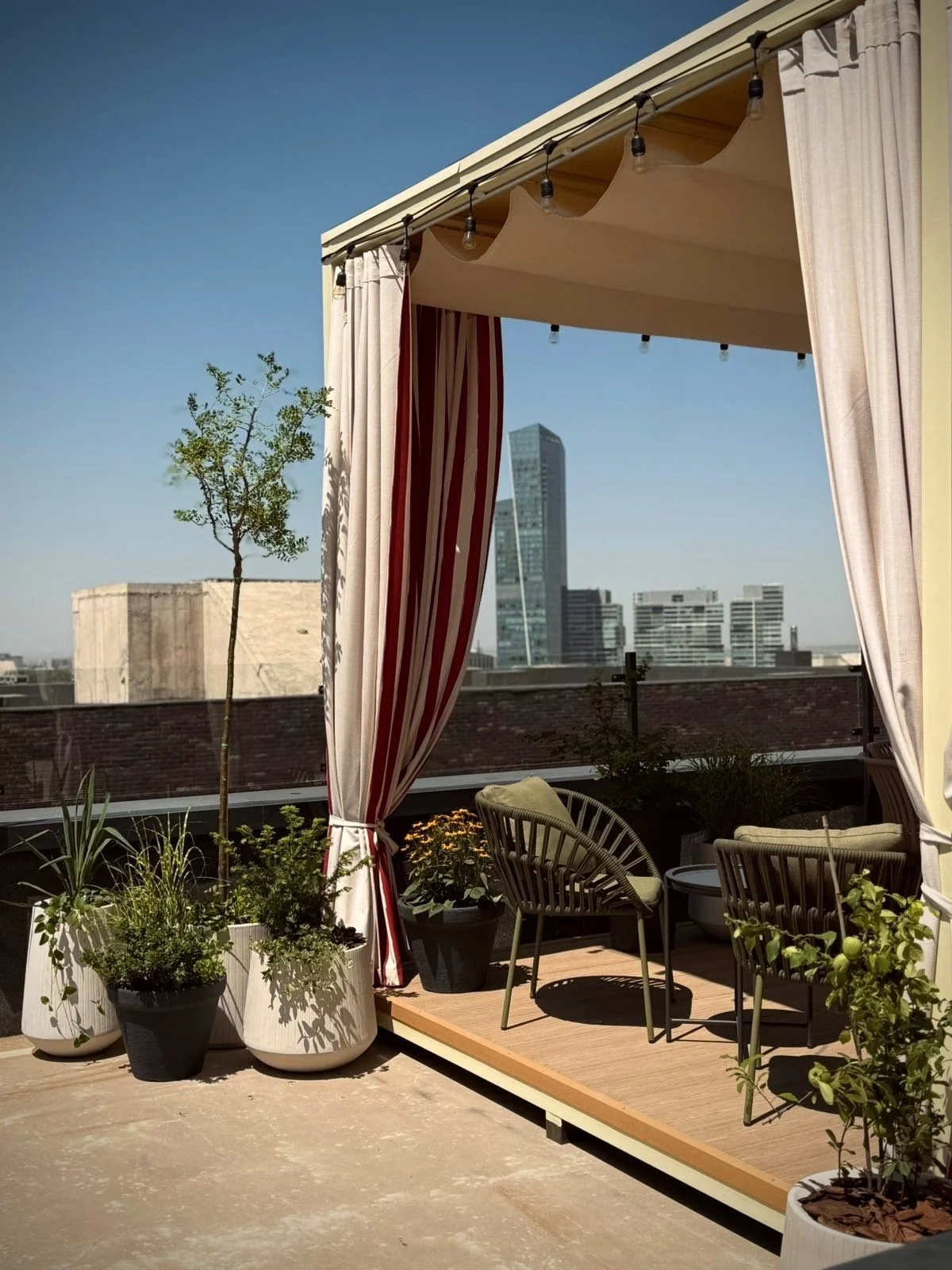 Rooftop terrace with potted plants, two chairs with cushions, and a canopy with curtains, overlooking a city skyline with modern buildings and a clear blue sky.