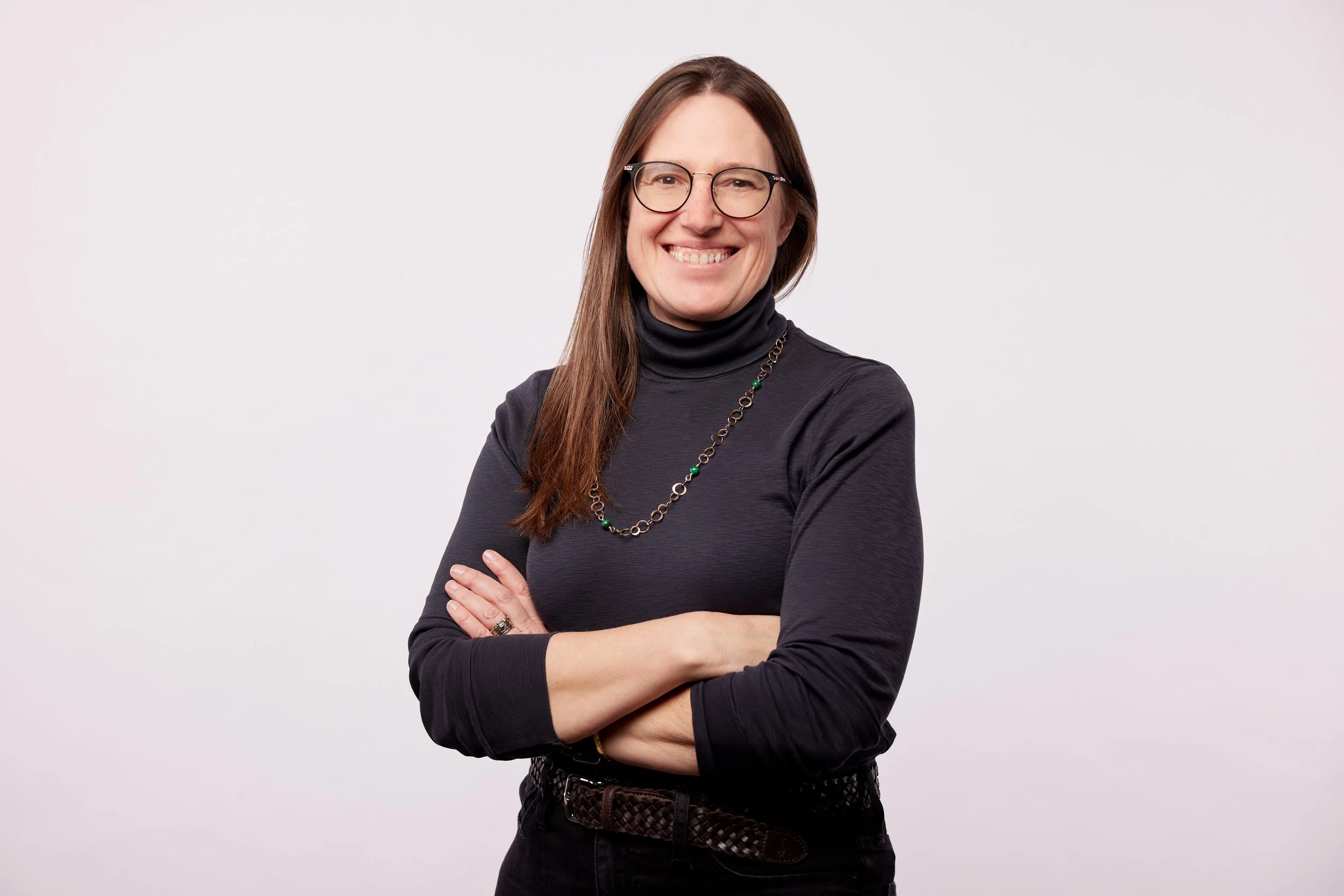 A woman with glasses and long brown hair smiling with arms crossed, wearing a black turtleneck and necklace against a plain white background.