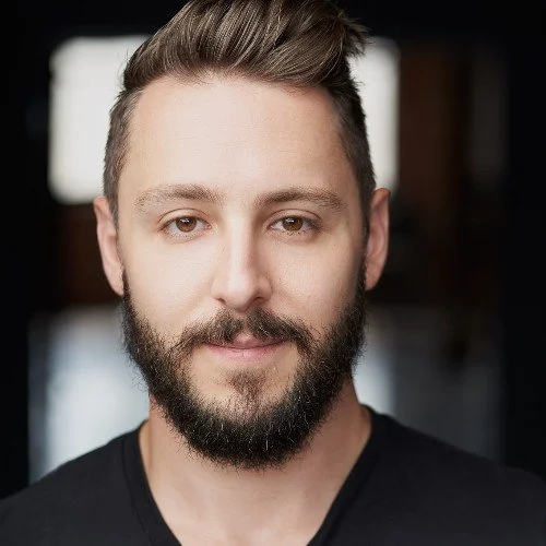 Close-up portrait of a young man with light skin, short hair, a beard, and a mustache, wearing a black shirt, looking at the camera with a slight smile in an indoor setting.