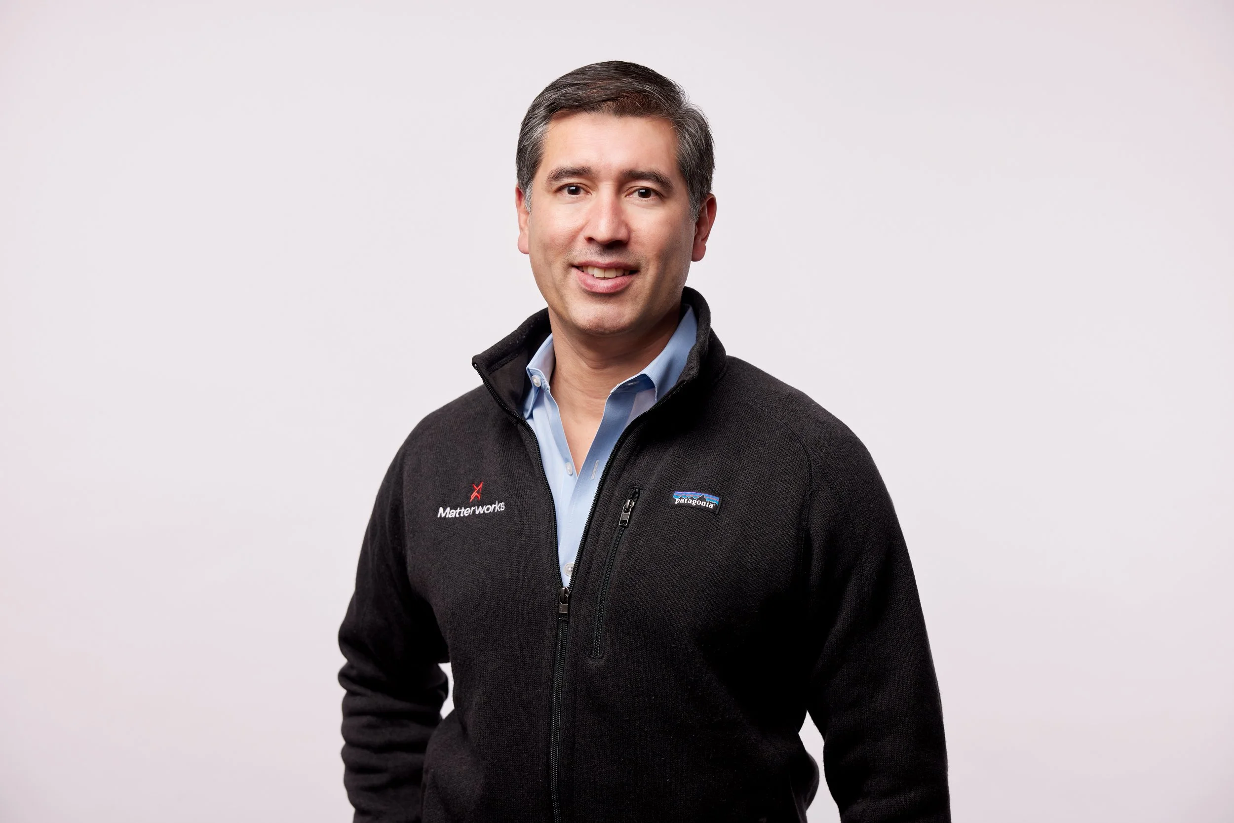 A man with dark hair smiling, wearing a black Patagonia fleece jacket over a light blue button-down shirt, standing against a plain light background.