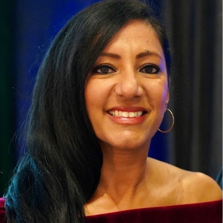 A smiling woman with long black hair, wearing makeup and gold hoop earrings, inside a venue with dark curtains.