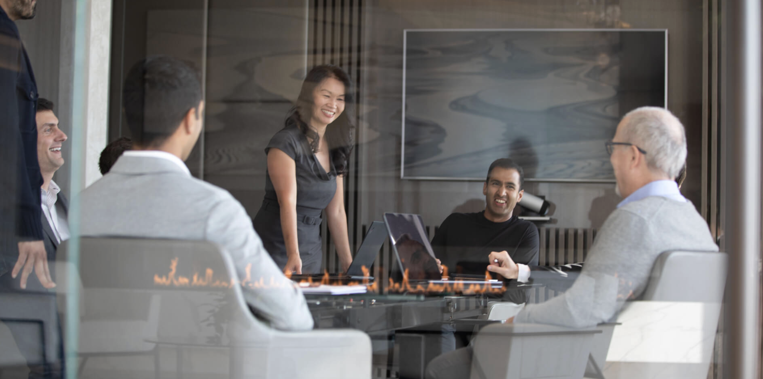 A group of five diverse professionals in a business meeting, smiling and engaging, with a woman standing and speaking at the center of a glass-walled conference room.