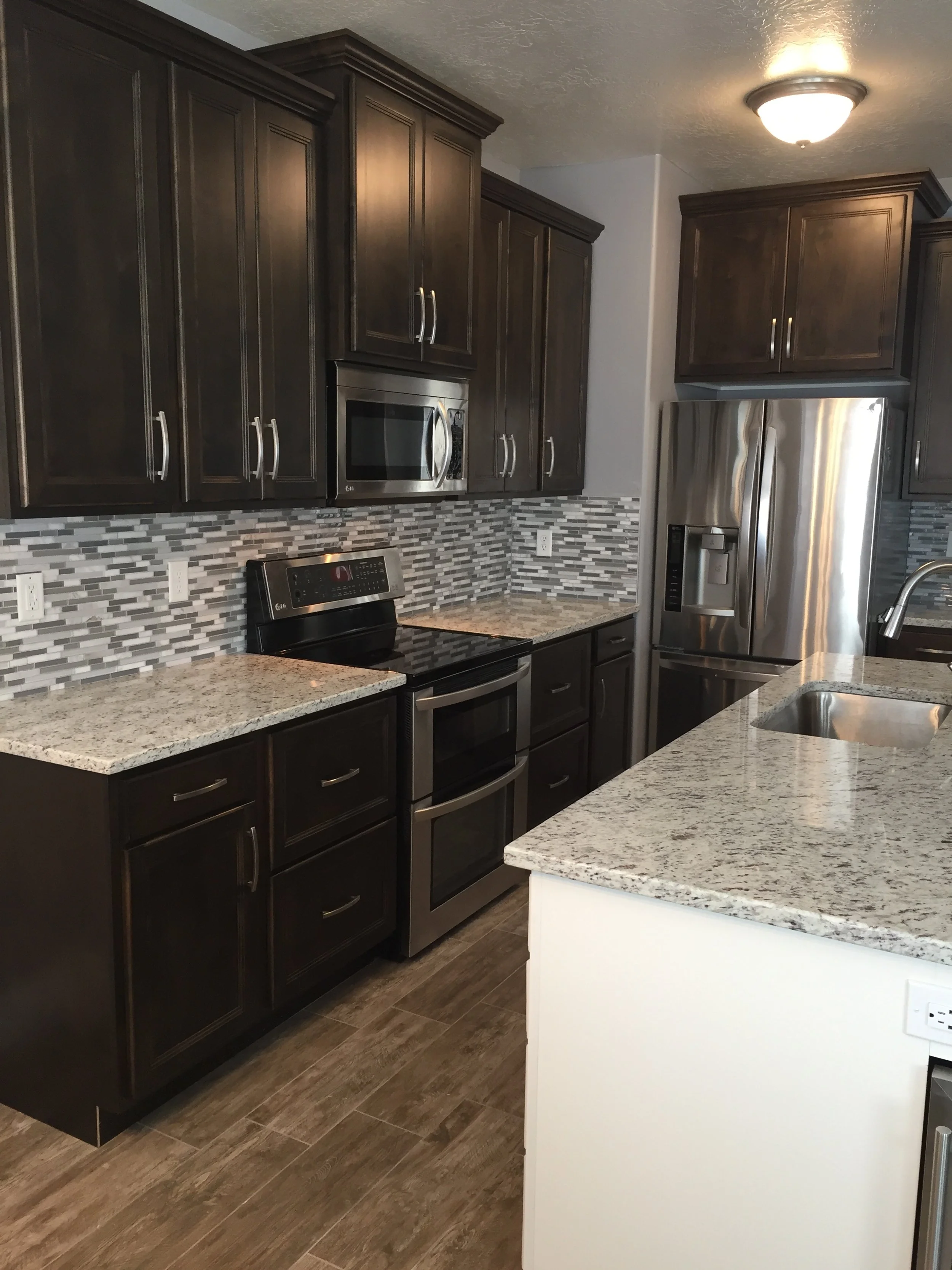Kitchen with dark wood cabinets, stainless steel refrigerator, microwave, and oven, granite countertops, mosaic tile backsplash, and wood-look flooring.