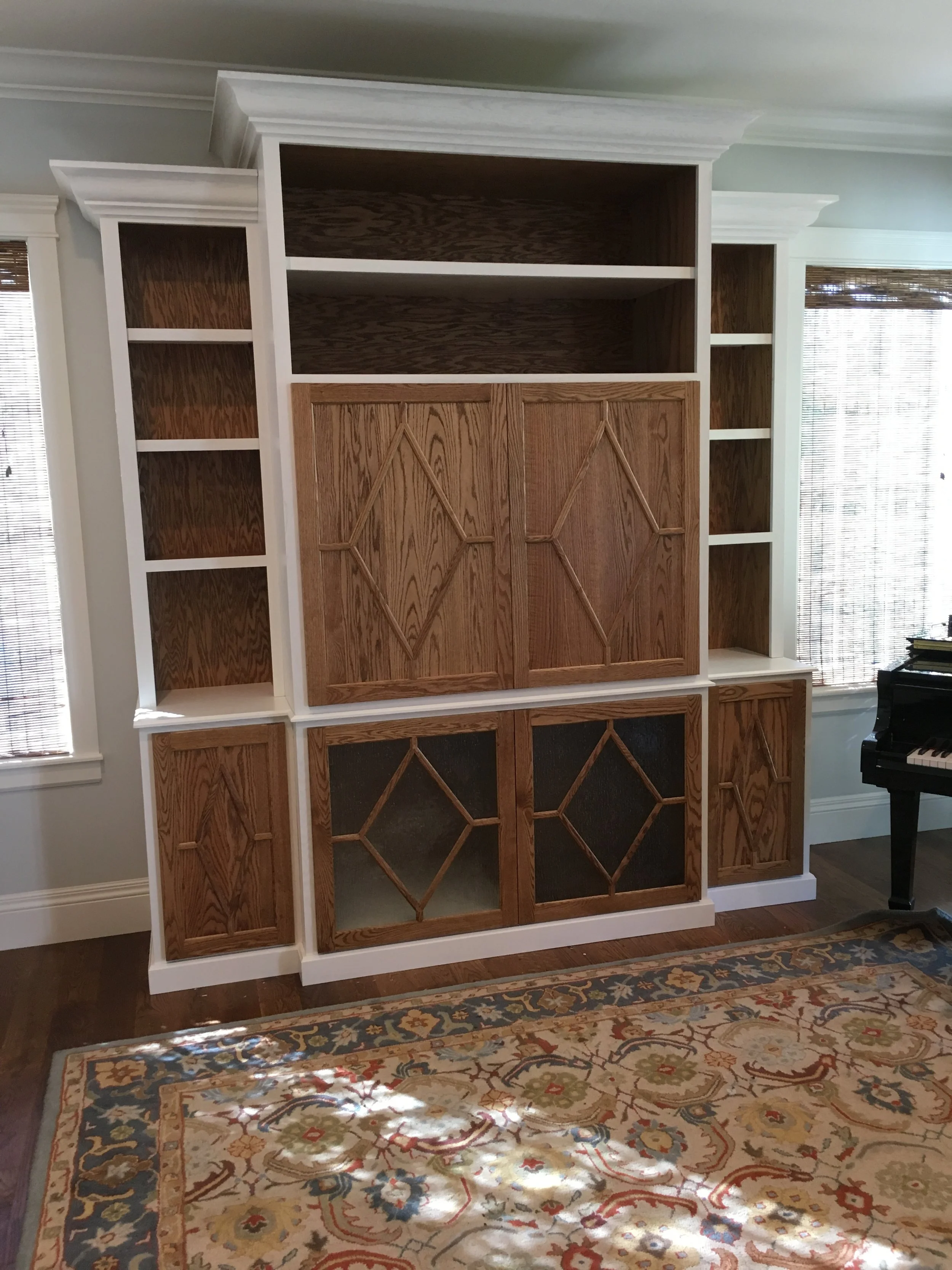 Empty built-in bookshelf with white and wood finishes, situated on a hardwood floor with an ornate area rug in front. Windows with blinds are on either side, and a black grand piano is partially visible on the right.
