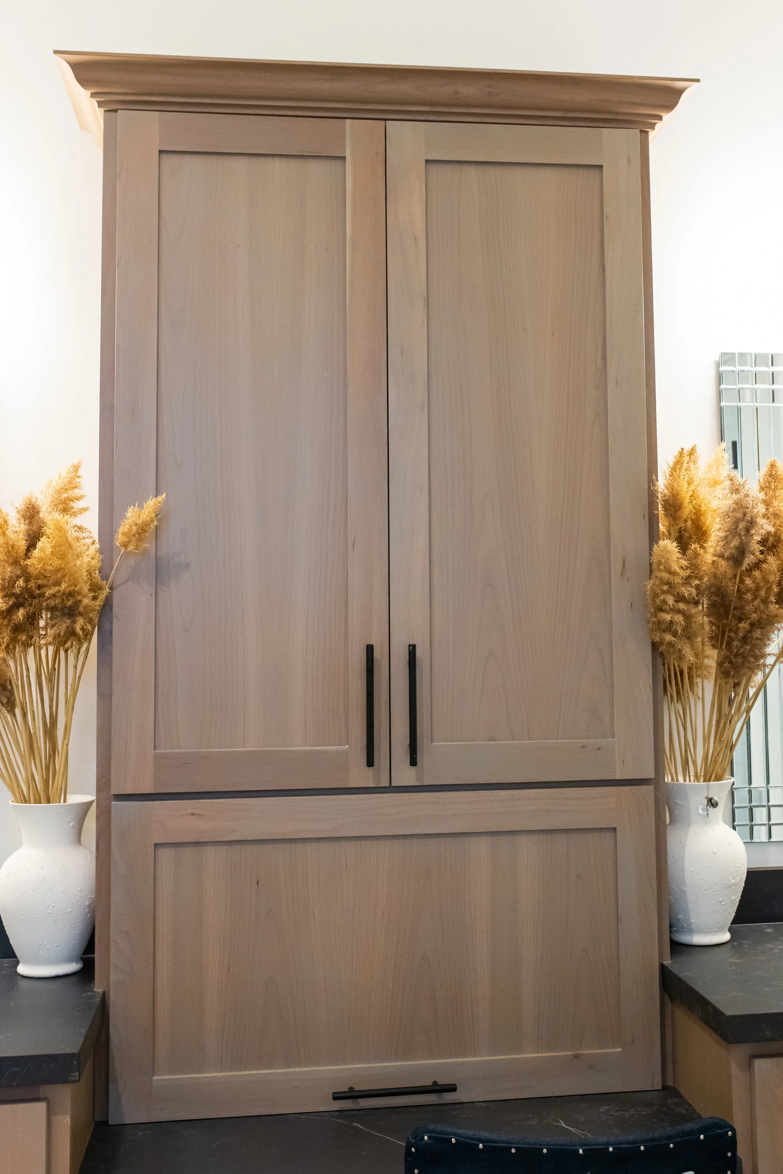 A large light wood wardrobe with black handles, flanked by two white vases with beige pampas grass and situated in a modern interior.